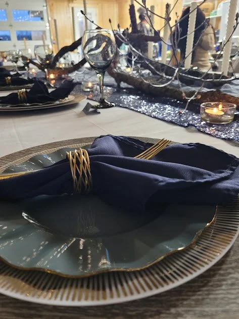 Elegant table setting with a gold-rimmed plate, navy napkin wrapped with gold-colored napkin rings, and a decorative centerpiece with branches, candles, and a glass goblet in the background.