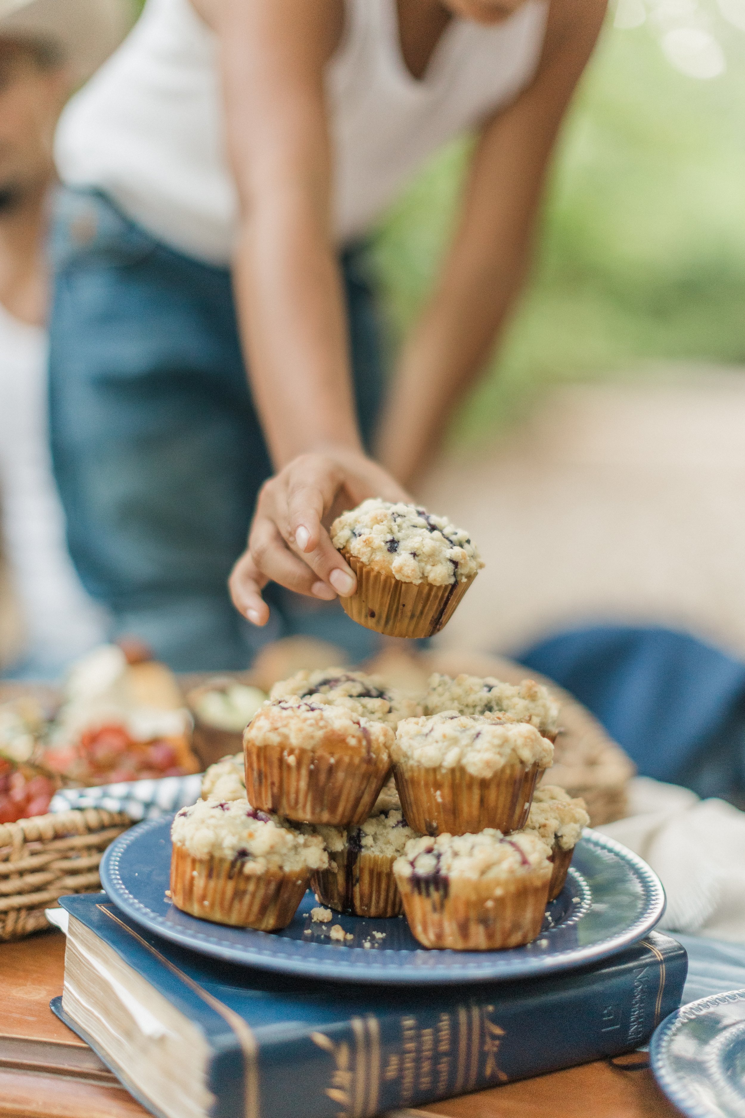 Person reaching for blueberry muffins on a blue plate at a gathering or outdoor event.