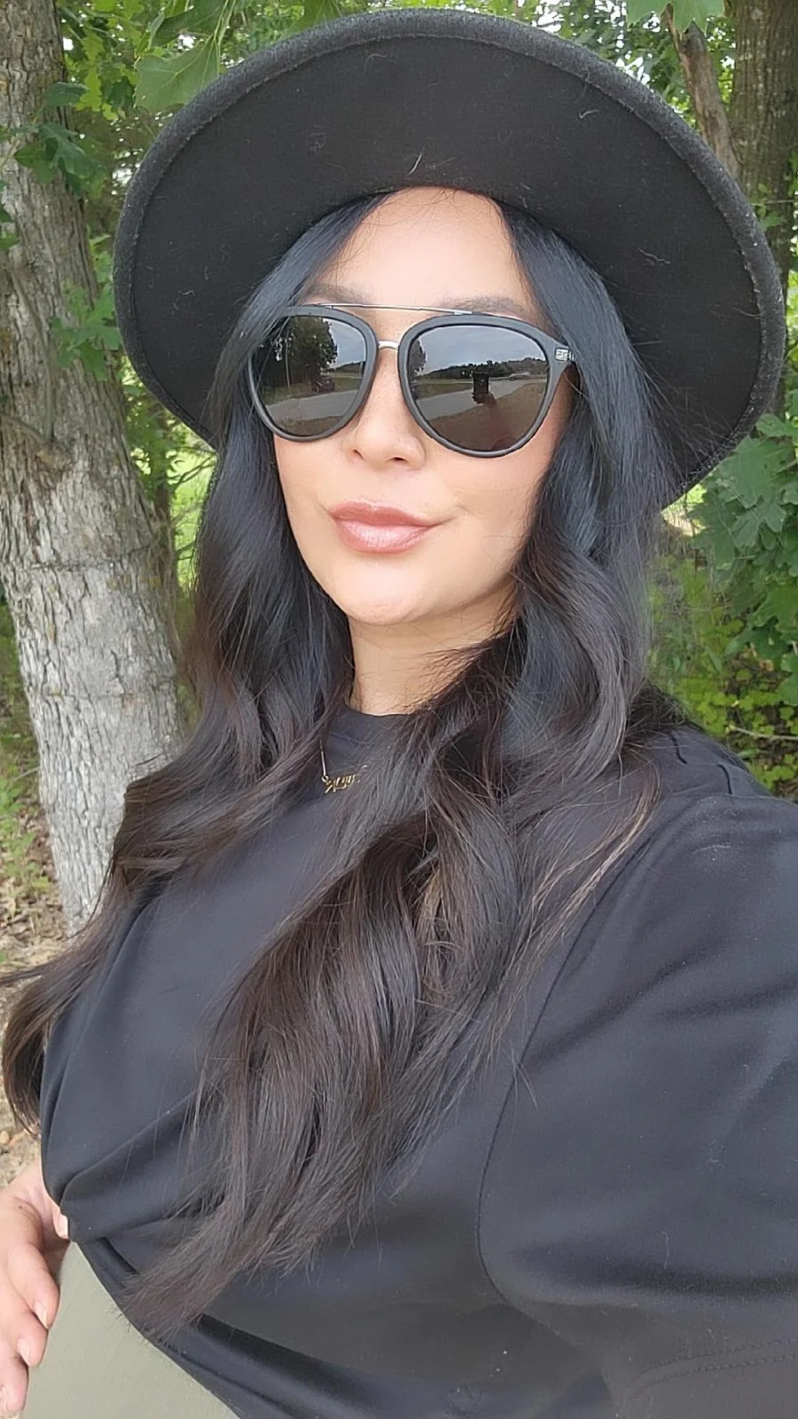 A woman with long wavy black hair wearing a black hat, large sunglasses, and a black top, standing outdoors near a tree with green leaves.