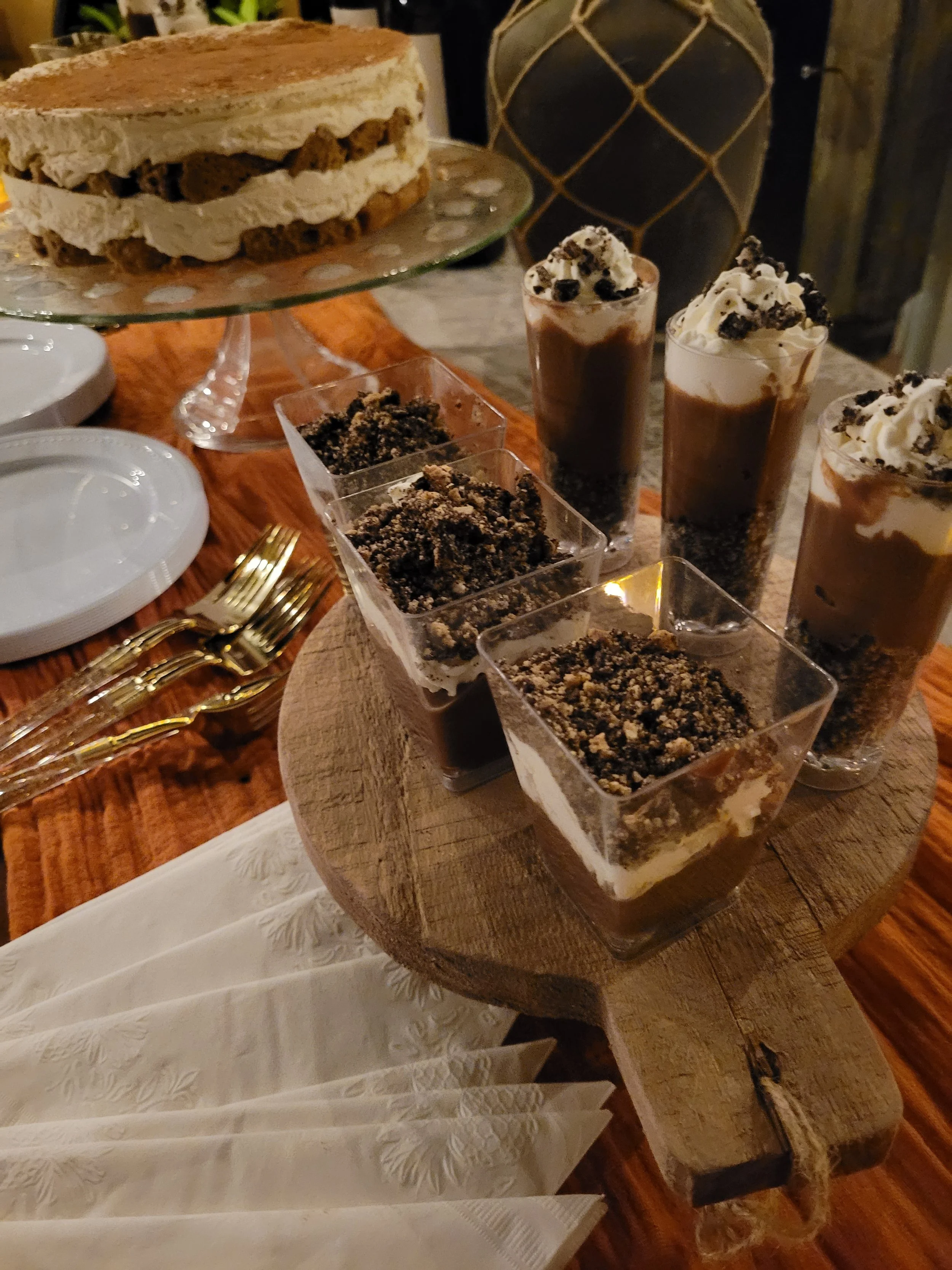 A table with a layered cake on a glass cake stand, glasses of chocolate mousse topped with whipped cream and cookie crumbs, and small square cups of crumbled cookies or dessert, all arranged on a rustic wooden serving board, with gold forks and plate