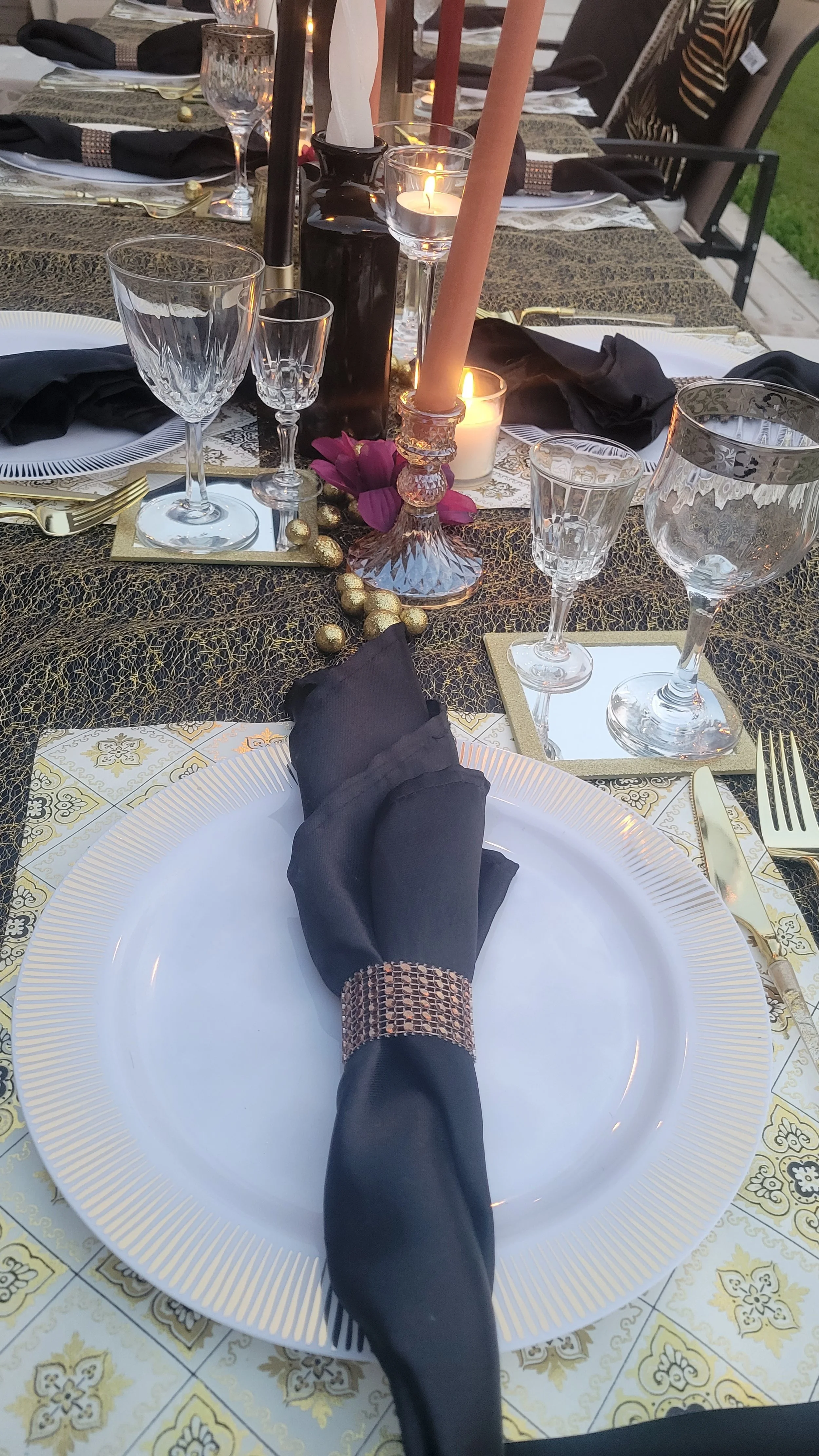 Elegant dinner table setup with black napkins, gold cutlery, crystal glasses, candle holders, and decorative gold accents.