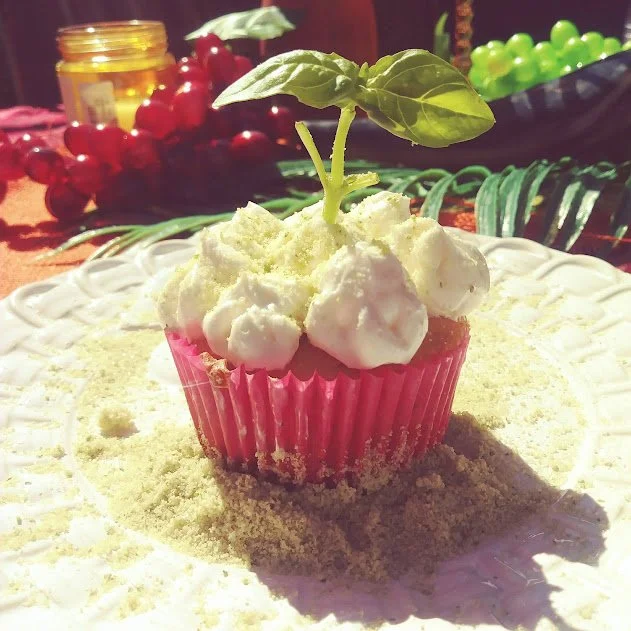 A cupcake with white frosting and a basil leaf on top, garnished with a green sprinkle, set on a plate with yellow powder around it, in a colorful background with red grapes and green grapes.