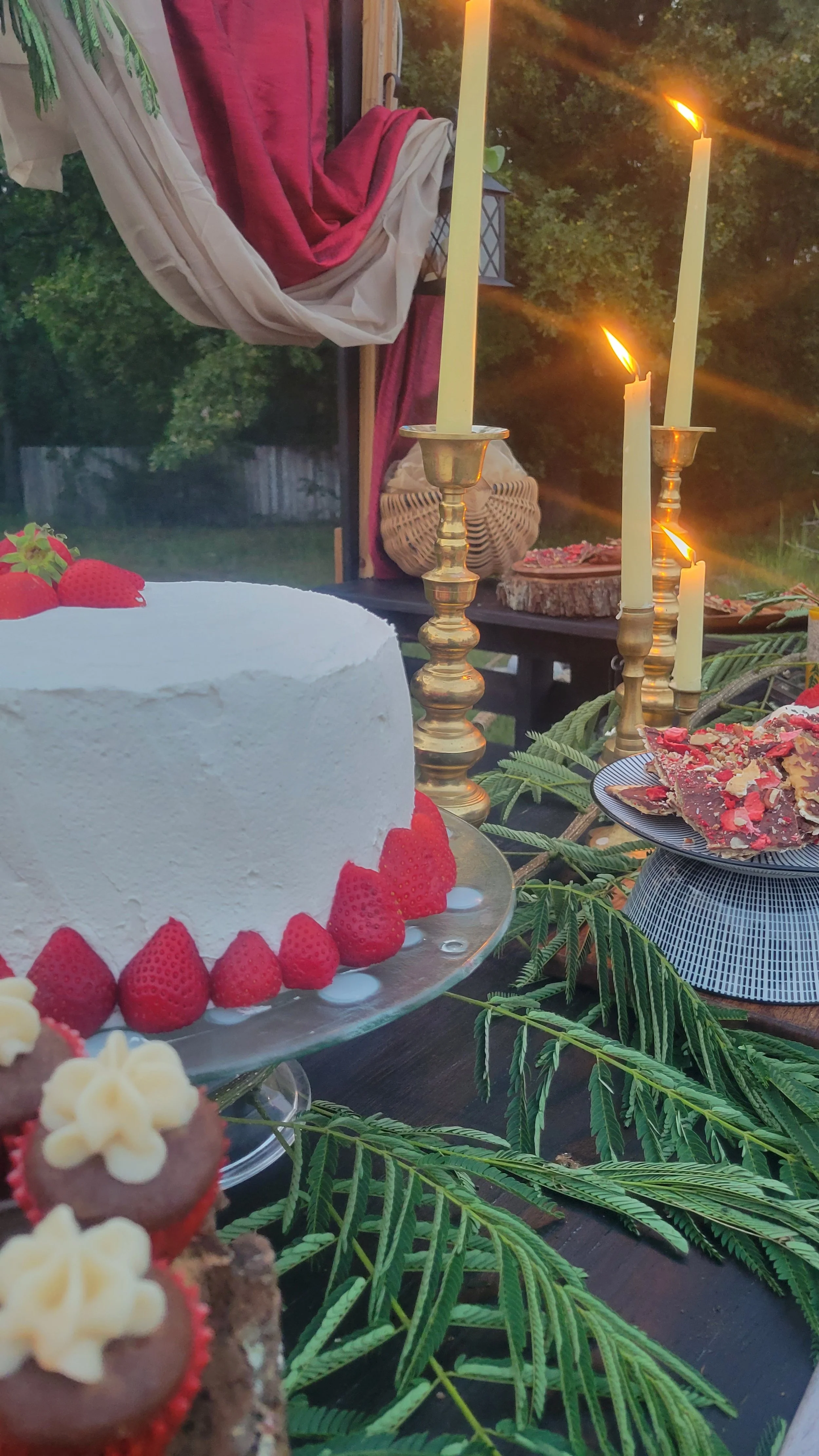 A table set outdoors with a white frosted cake decorated with strawberries, various chocolates, and flowers, surrounded by green leaves, lit candles in gold candlesticks, and a background of trees and sunset.