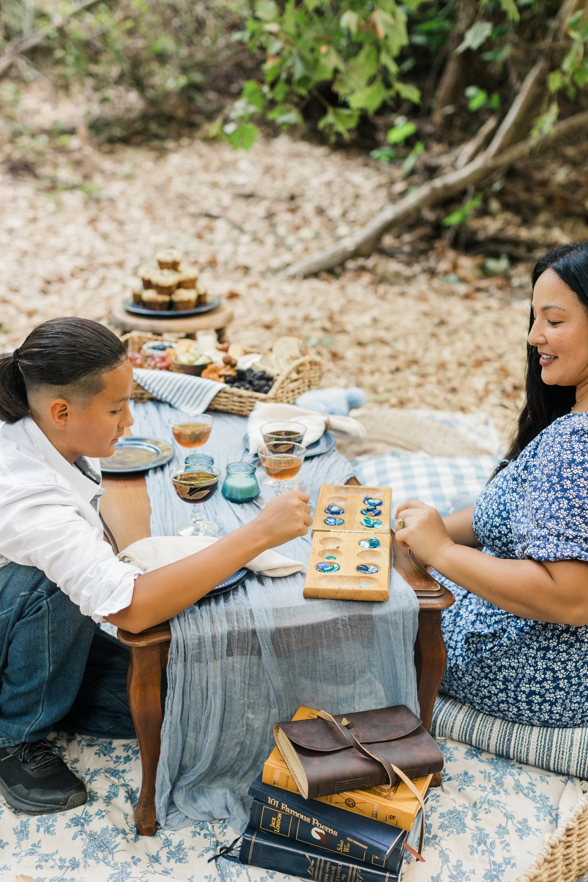Two women having a tea party outdoors, playing with glass marbles at a low wooden table, with food and drinks, featuring stacked cookies, on a picnic blanket surrounded by books and natural setting.