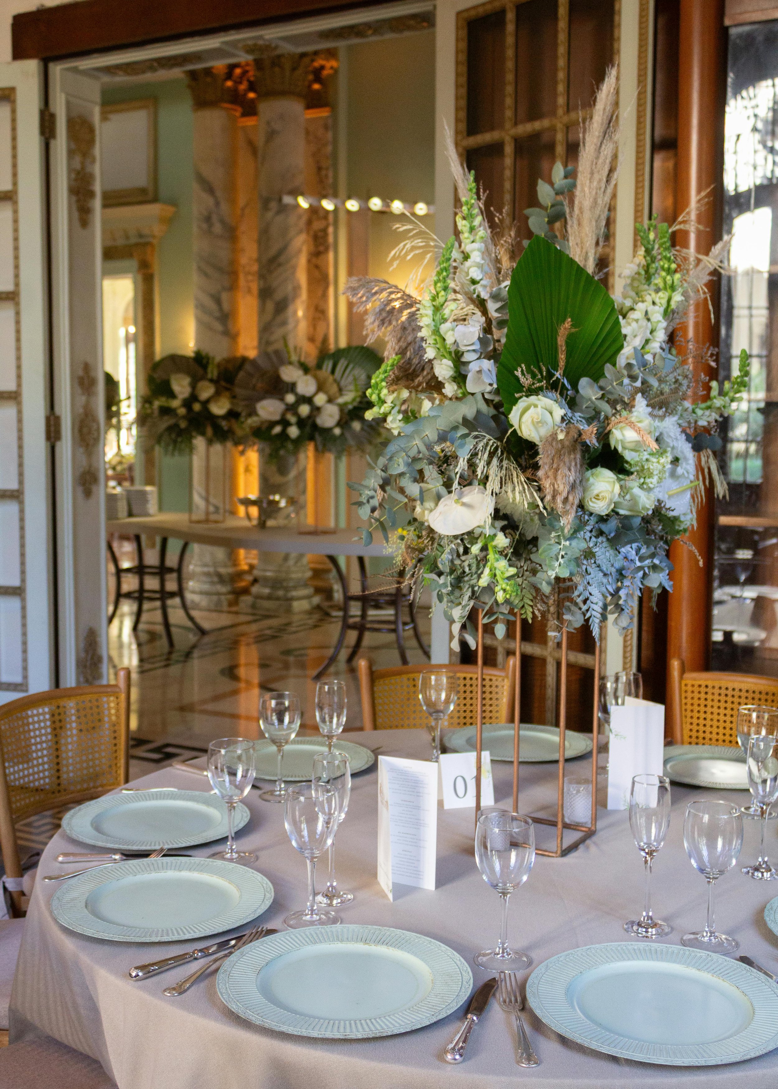 A formal dining table set with white plates, silverware, wine glasses, and a large floral centerpiece in a decorated elegant room.