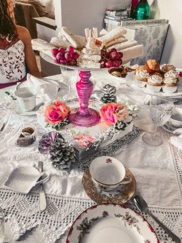 A decorated table with a pink candle holder, pink and purple flowers, pine cones, and assorted desserts including cookies and cupcakes, set with white lace tablecloth and fine china.