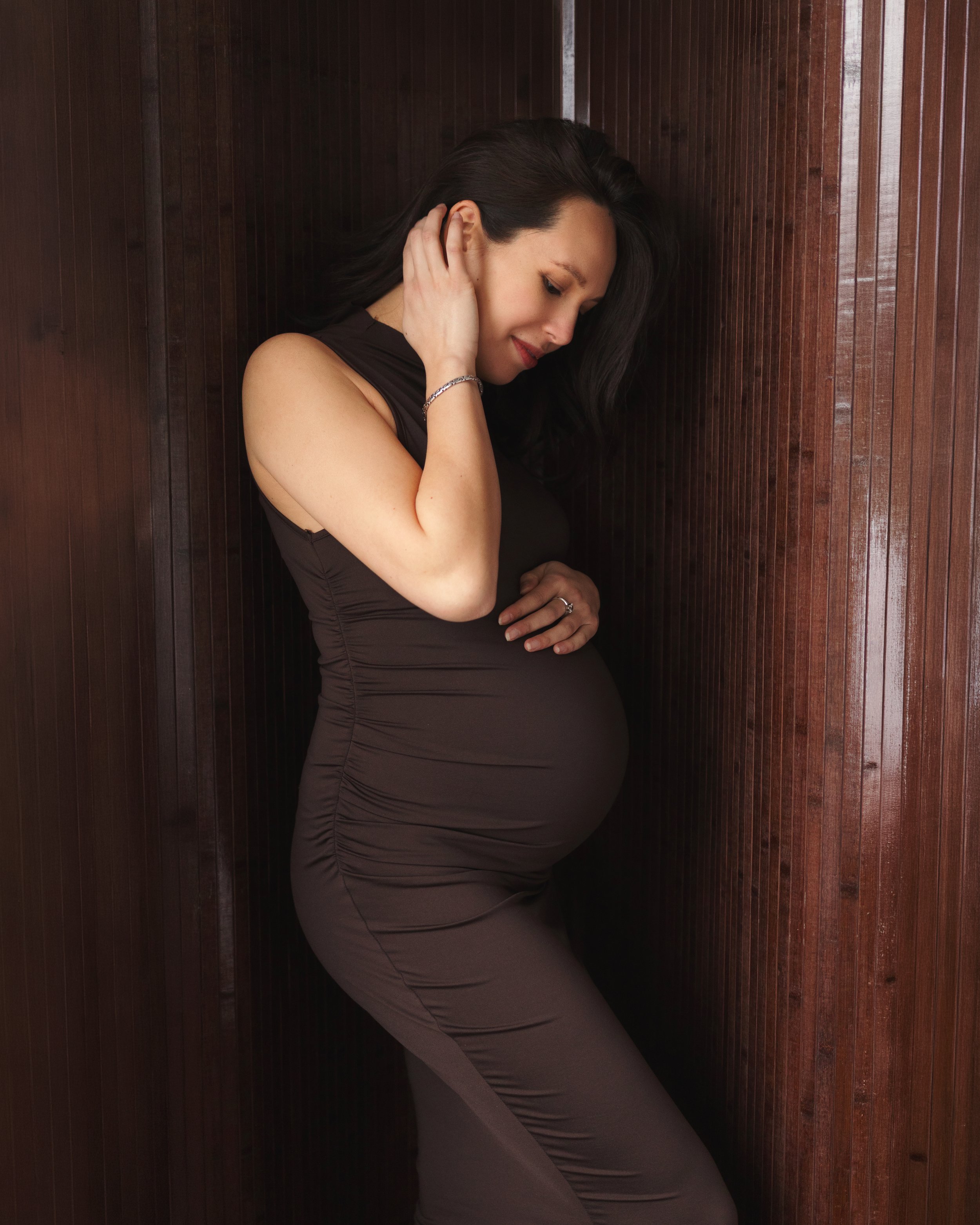 Pregnant woman in brown outfits leaning against a wooden wall, softly smiling with hand touching her head.