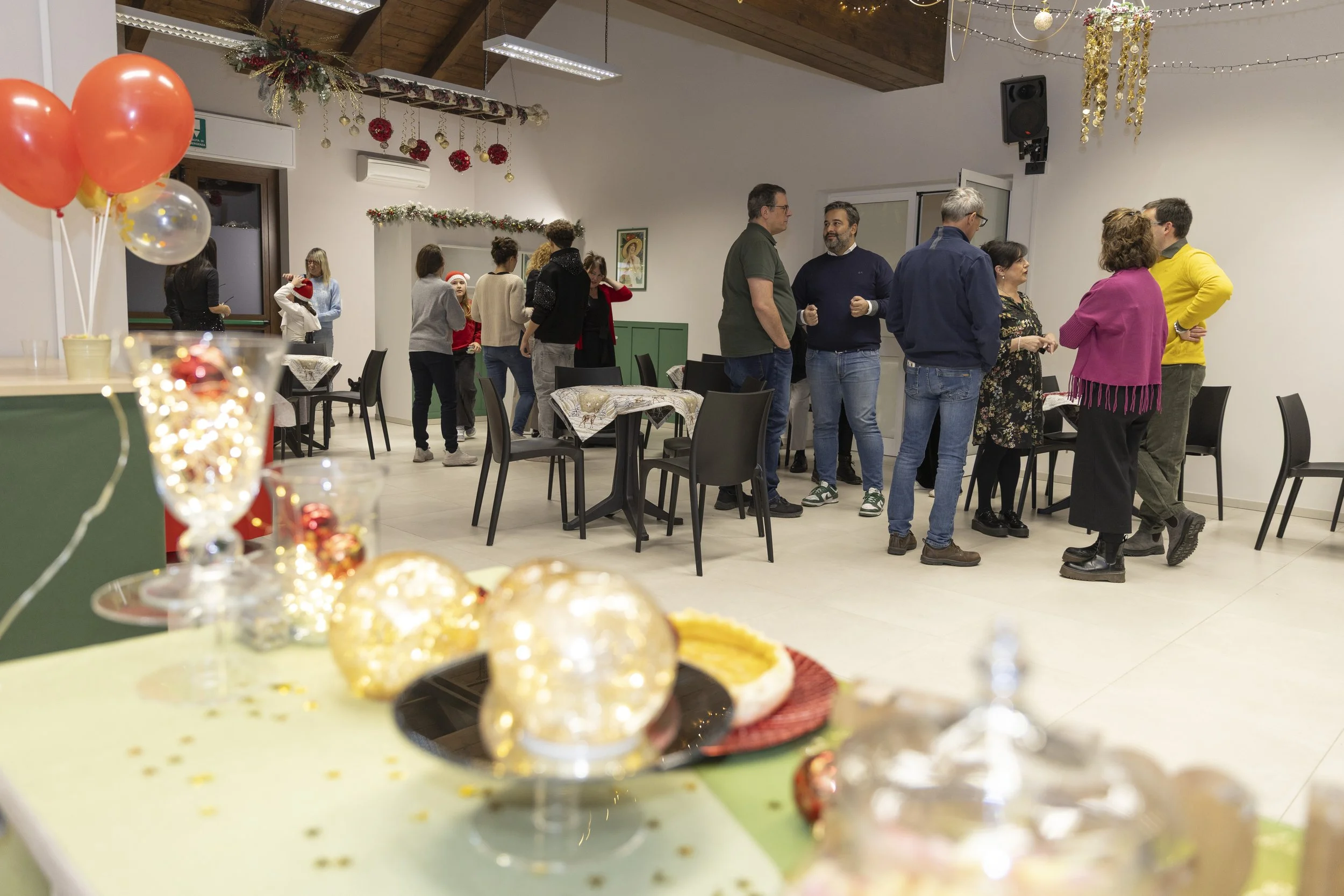 Festa di Natale con persone che socializzano, decorazioni natalizie come palline e luci, in un ambiente spazioso con tavoli e sedie.