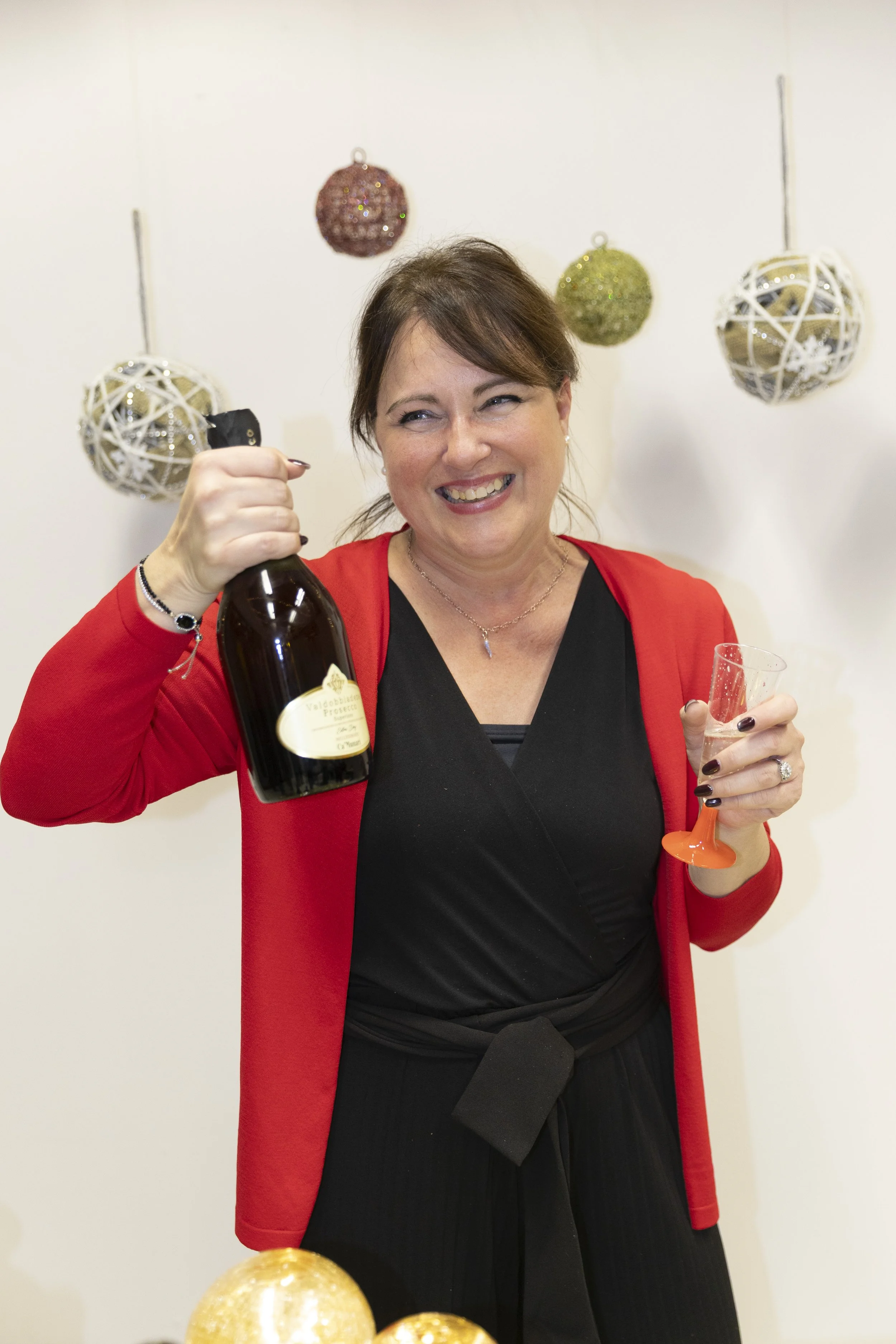Una donna sorridente con un vestito nero e una giacca rossa tiene una bottiglia di champagne e un bicchiere vuoto, celebrando un evento festivo. Sullo sfondo decorazioni natalizie.