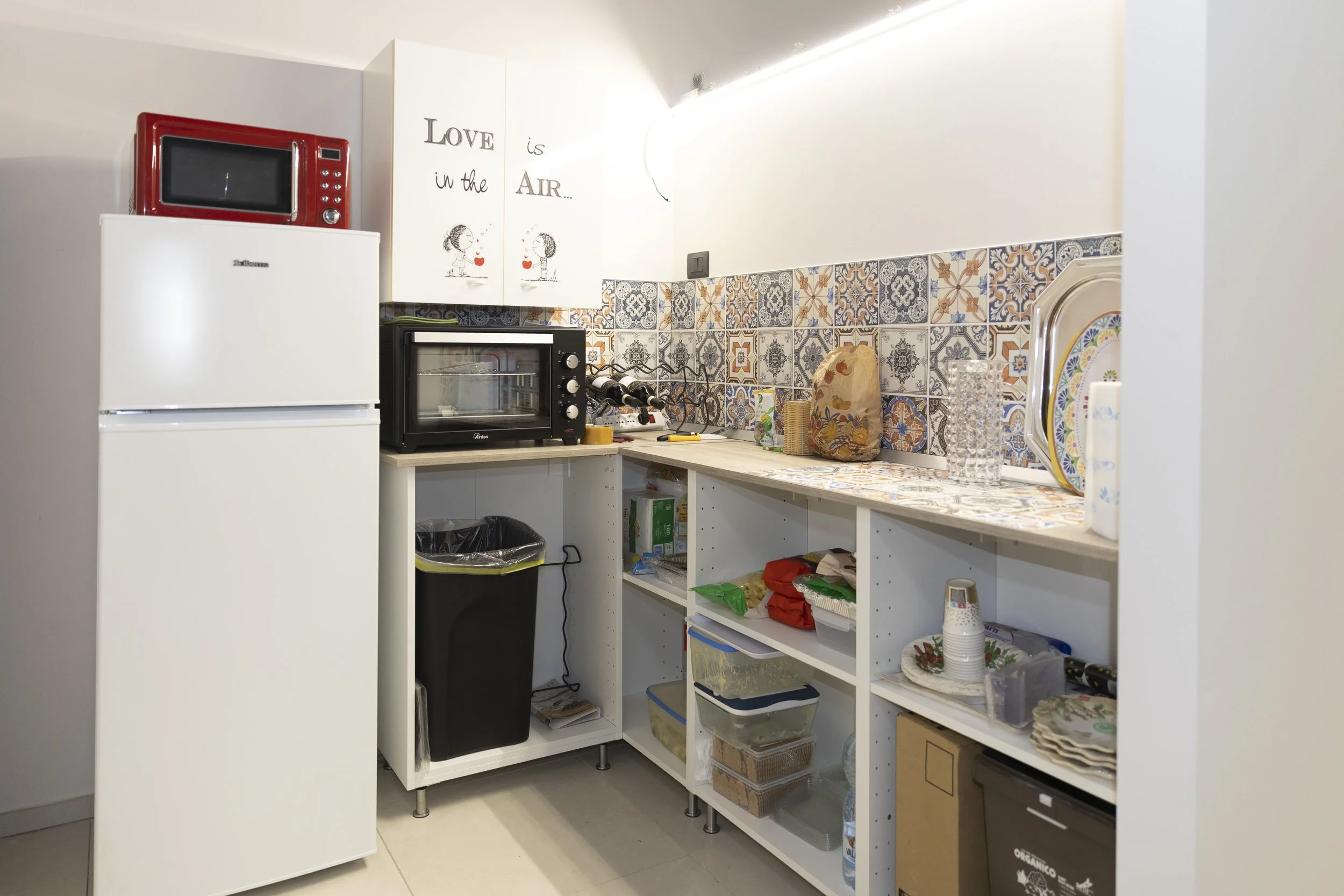 Una cucina con frigorifero bianco, forno piccolo, microonde rosso, scaffali con piatti, bicchieri e vari oggetti.