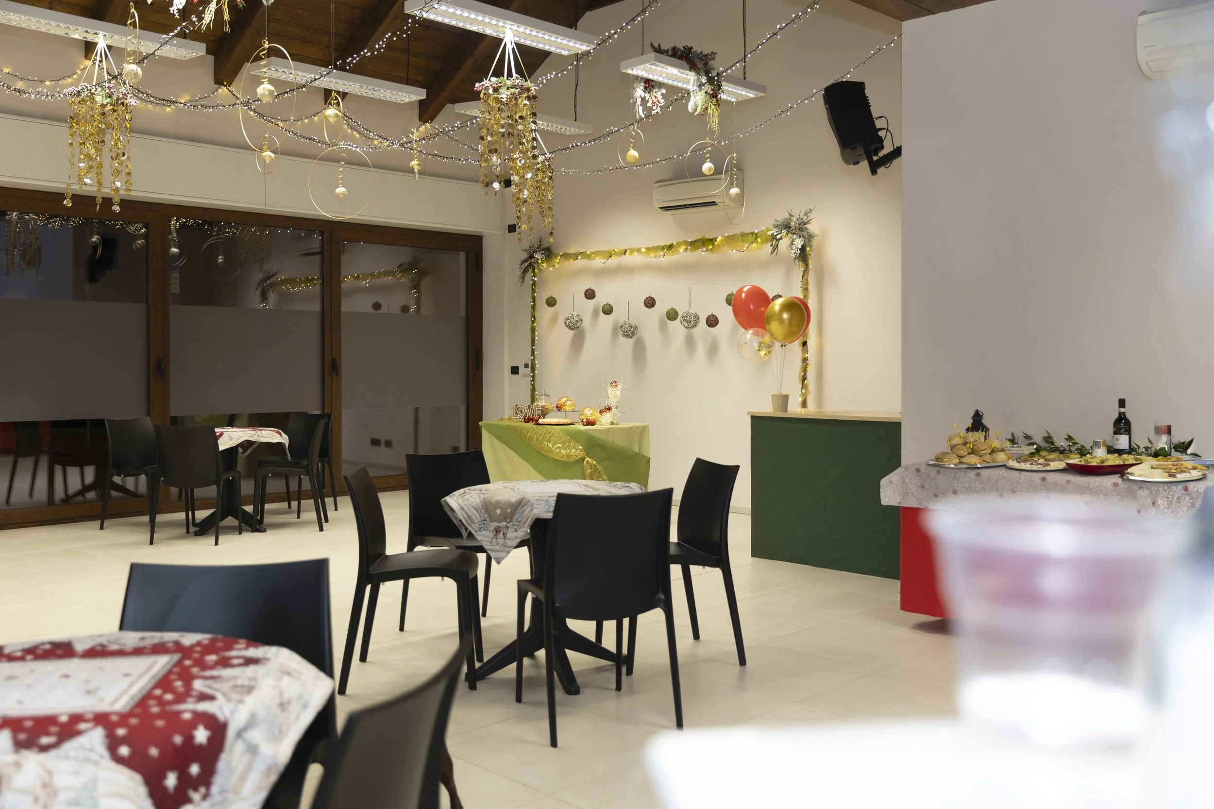 Sala decorata per una festa natalizia con luci, palloncini rossi e d'oro, e decorazioni natalizie alle pareti, con tavoli apparecchiati per un evento.