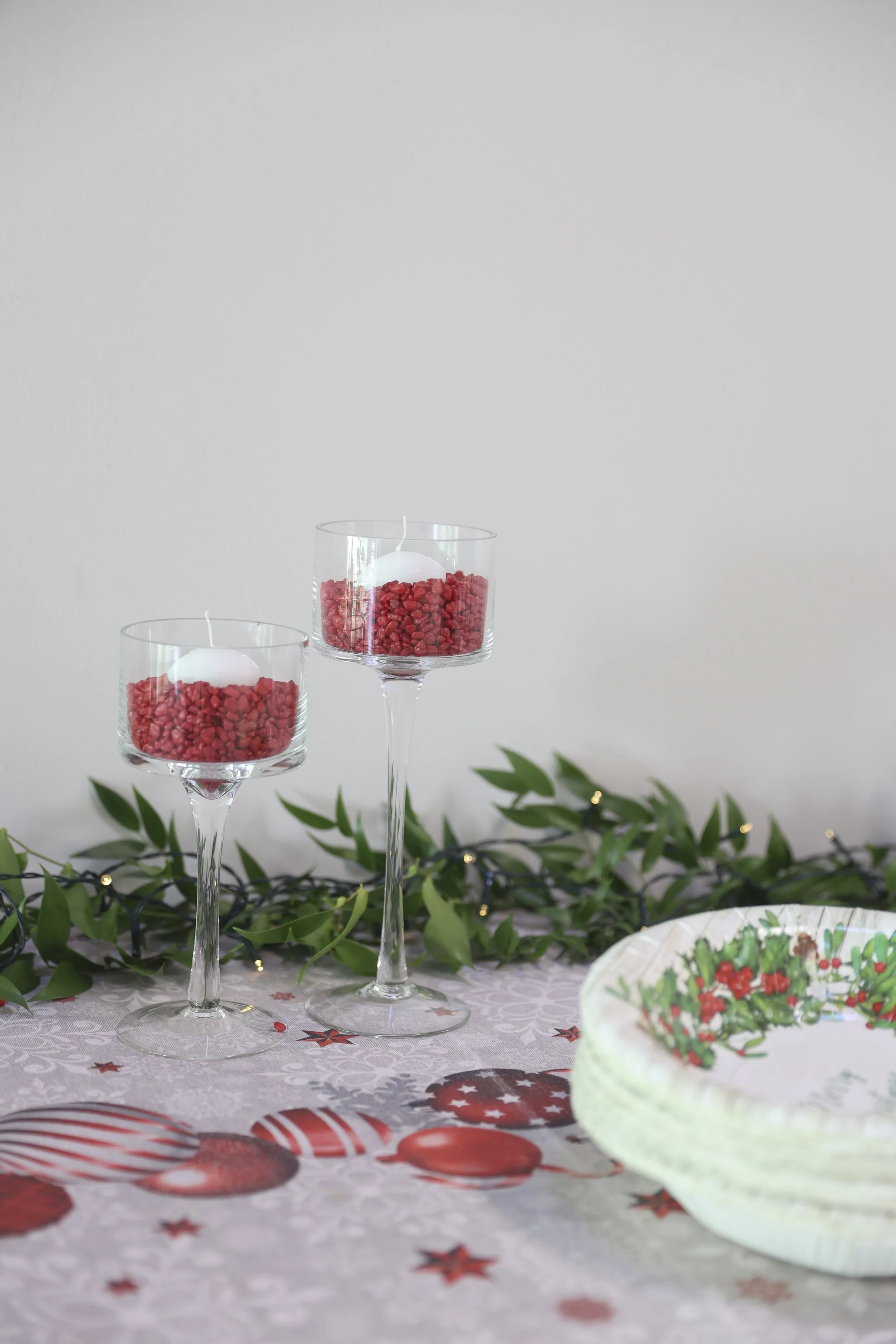 Due bicchieri di vetro con candele galleggianti in rivestimenti di ciottoli rossi, decorati con rami di piante verdi e luci natalizie, su un tavolo con tovaglia decorata con motivi natalizi e un piatto con decorazioni natalizie.