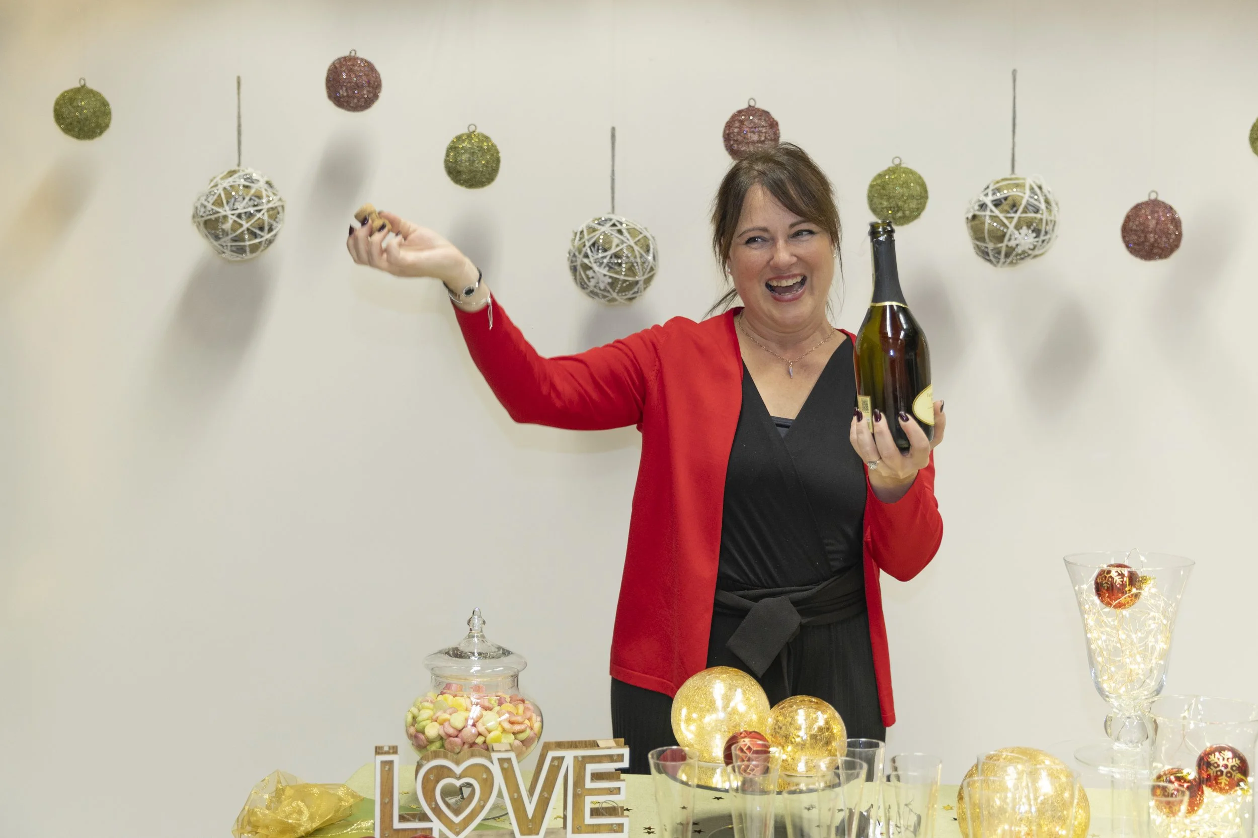 Donna felice con boccetta di spumante davanti a decorazioni natalizie, tra cui palline colorate e luci, e un' insalatiera con caramelle