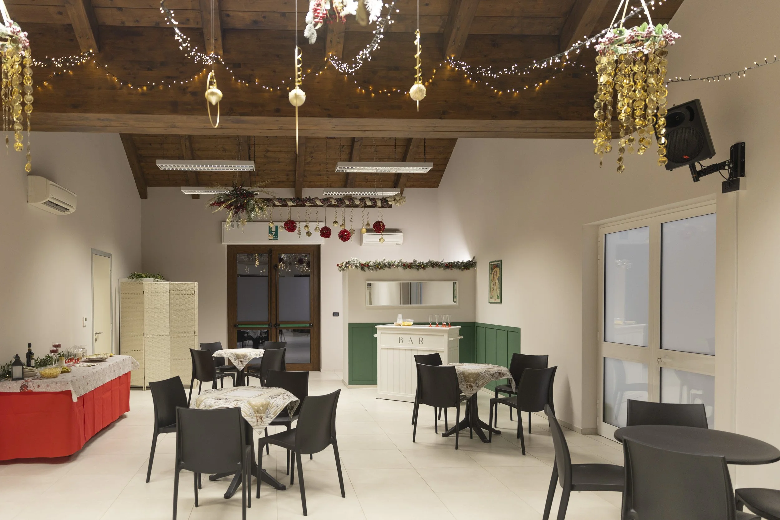 Sala decorata per una festa natalizia con tavoli, sedie, decorazioni di luci e ornamenti natalizi, bar con decorazioni festive e pareti con decorazioni natalizie.