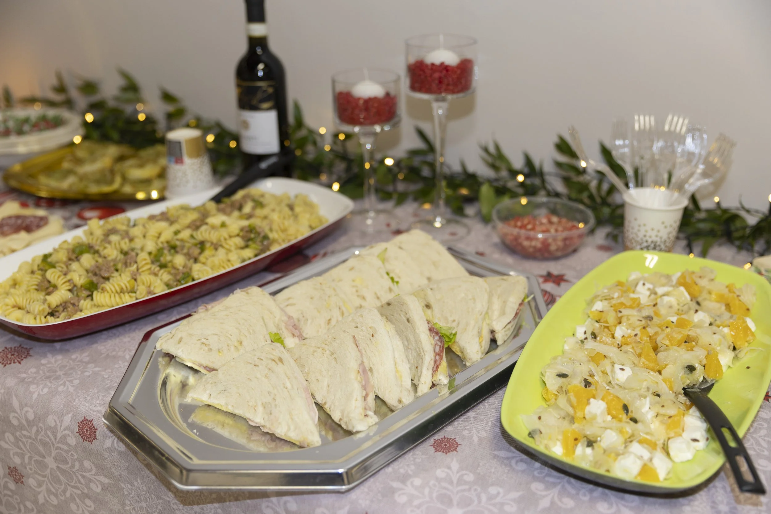 Sei piatti di cibo su un tavolo: un vassoio di panini, una teglia di pasta con carne, un piatto di insalata di frutta e due bicchieri con decorazioni di Natale. Sullo sfondo, una bottiglia di vino, bicchieri di vetro con decorazioni natalizie e luci 