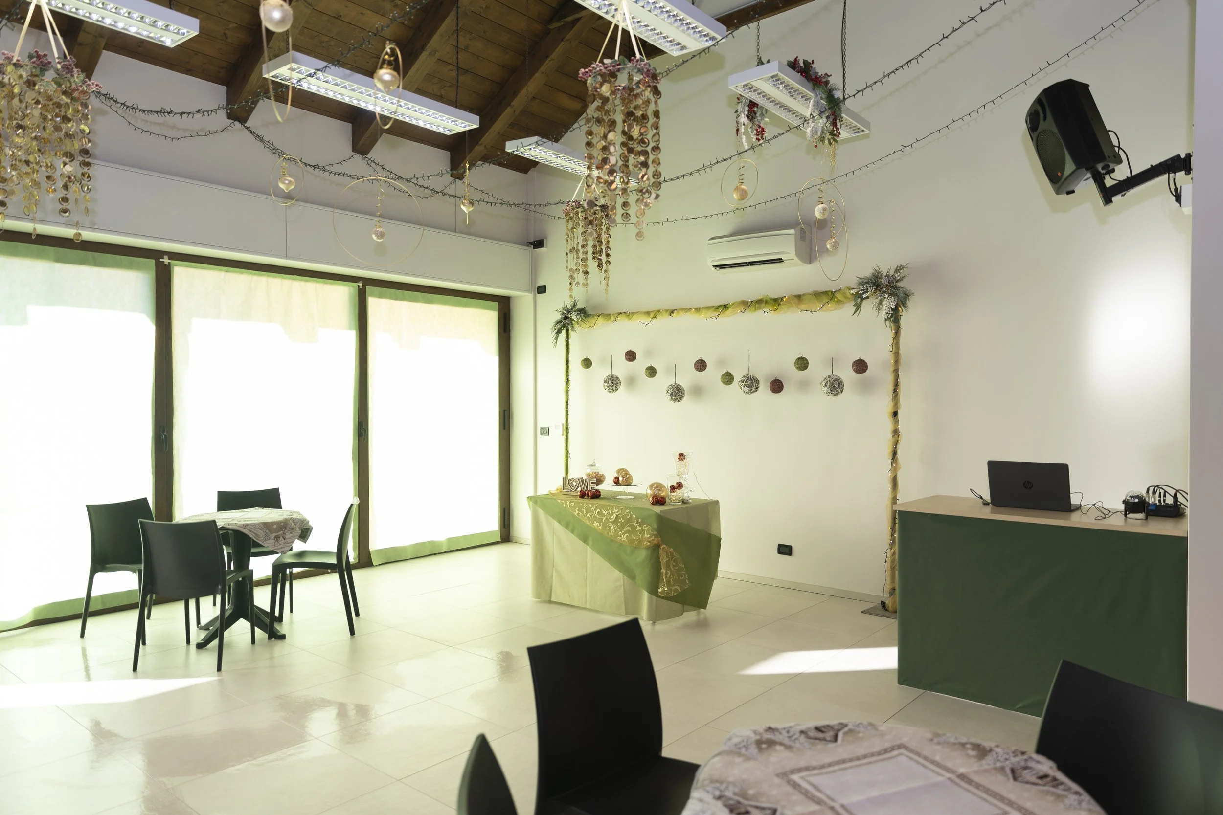 Stanza decorata con festoni natalizi, tavolo con decorazioni, sedie nere, pareti bianche, finestre con tende chiare, soffitto in legno con luci decorative, apparecchio di aria condizionata, pulcino decorativi e schermo audio.