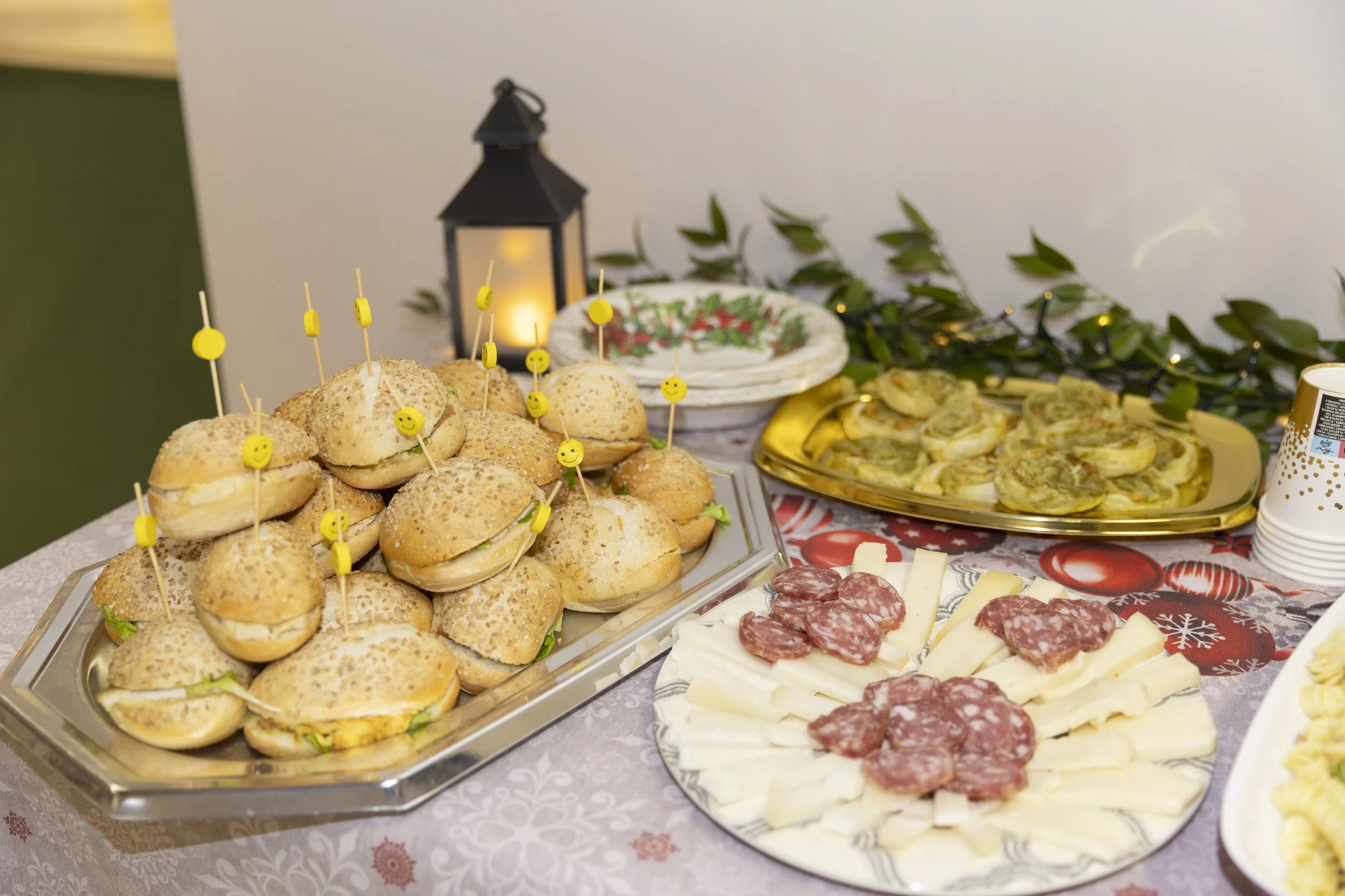 Antipasto con mini panini con sfilini di smiley, formaggi, salumi, e altri stuzzichini su un tavolo decorato con foglie verdi e luci natalizie.