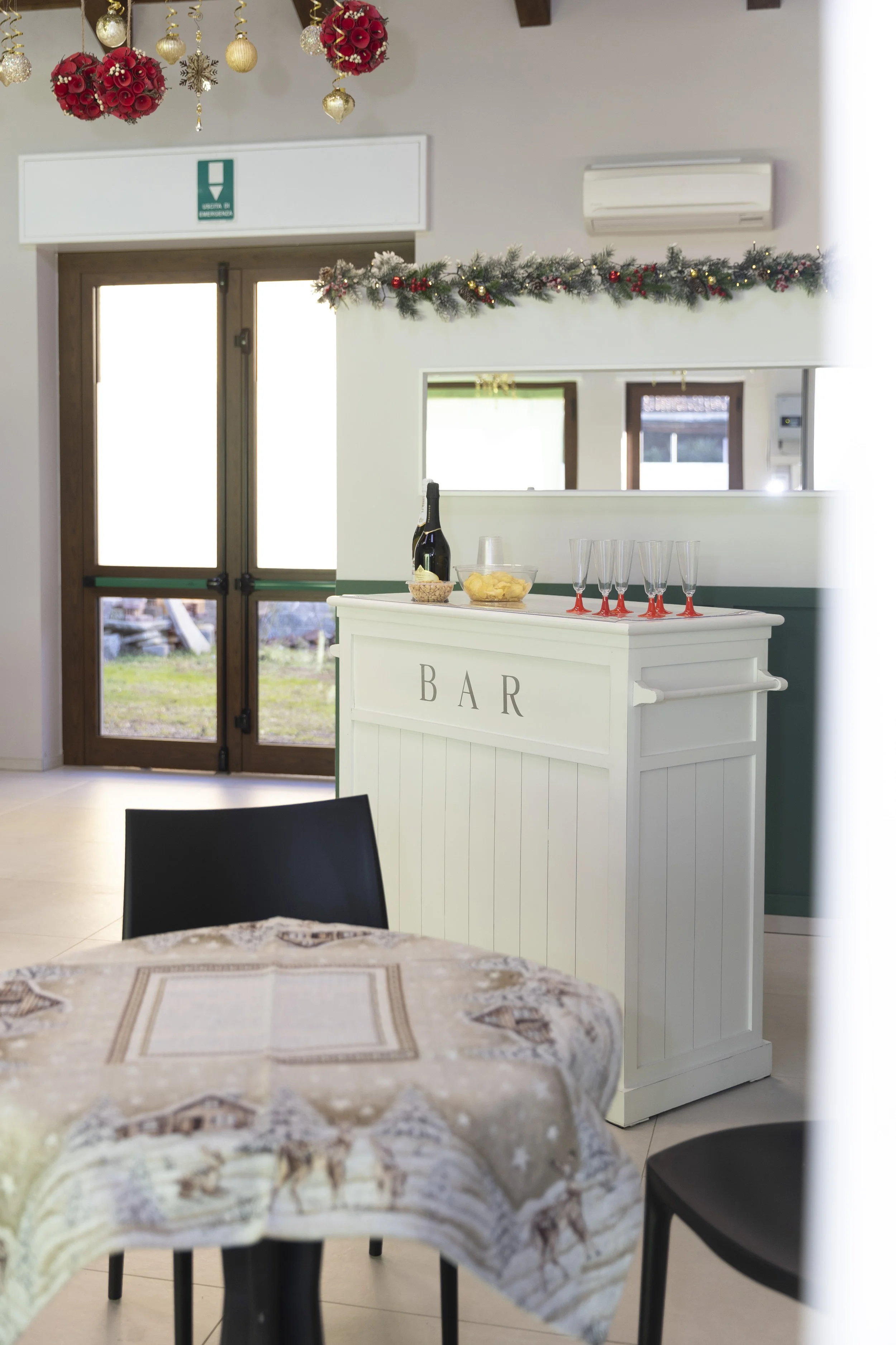 Interno di un bar decorato per le festività natalizie con addobbi a tema rosso e oro, un tavolo apparecchiato con una tovaglia natalizia, e una piccola zona bar con bottiglia di champagne e bicchieri.