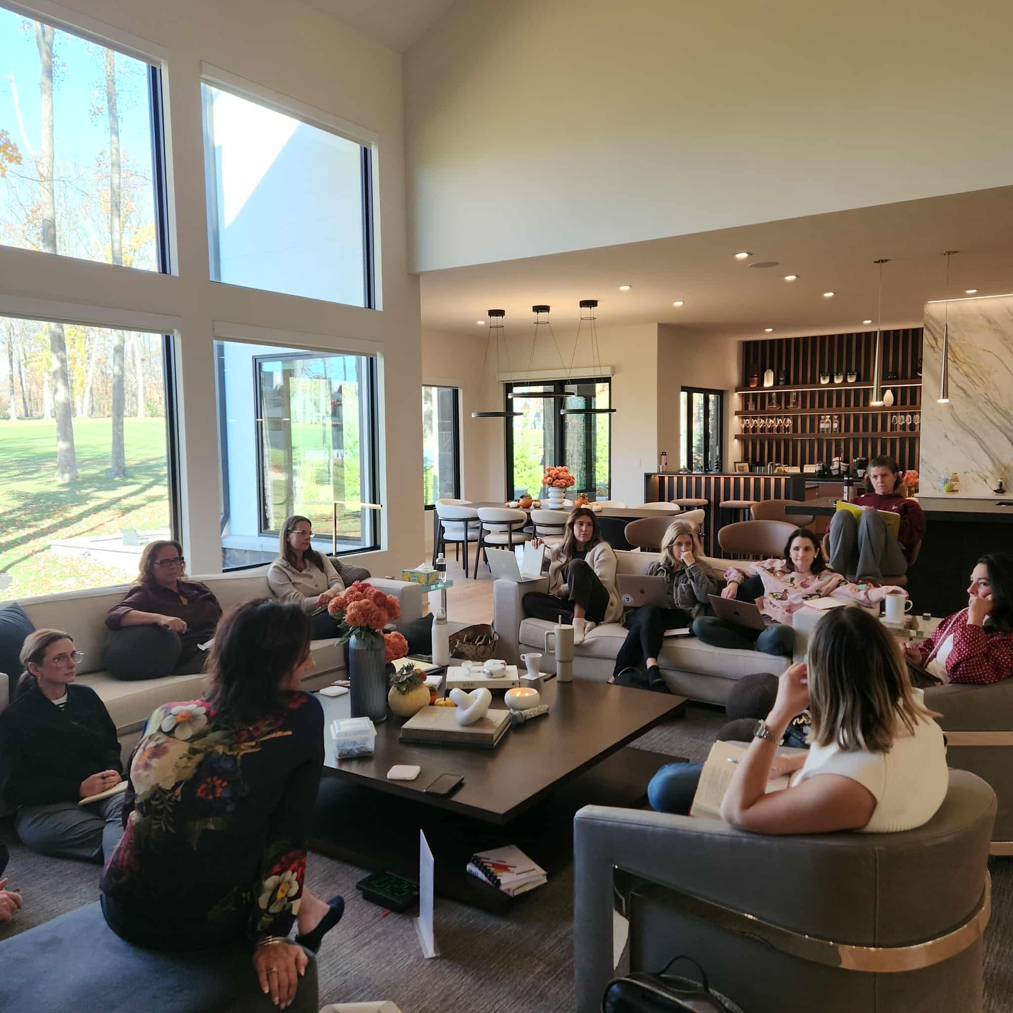 Collective members seated together during a group coaching session. A group of people sitting in a spacious, modern kitchen or dining area with large windows, enjoying a social gathering or meeting.