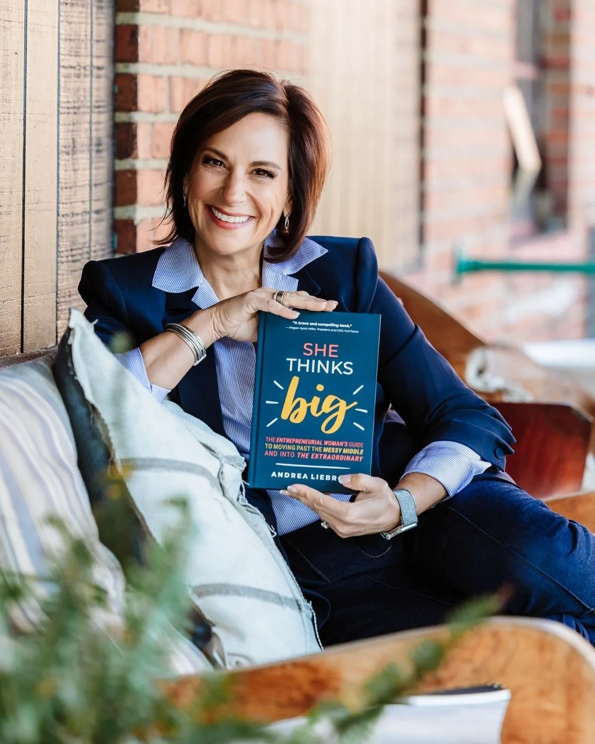 Andrea Liebross smiling in a navy blazer in a portrait shot. A woman with short brown hair, wearing a navy blazer and striped shirt, smiling and sitting on a porch, holding a book titled 'She Thinks Big' with a brick wall background.
