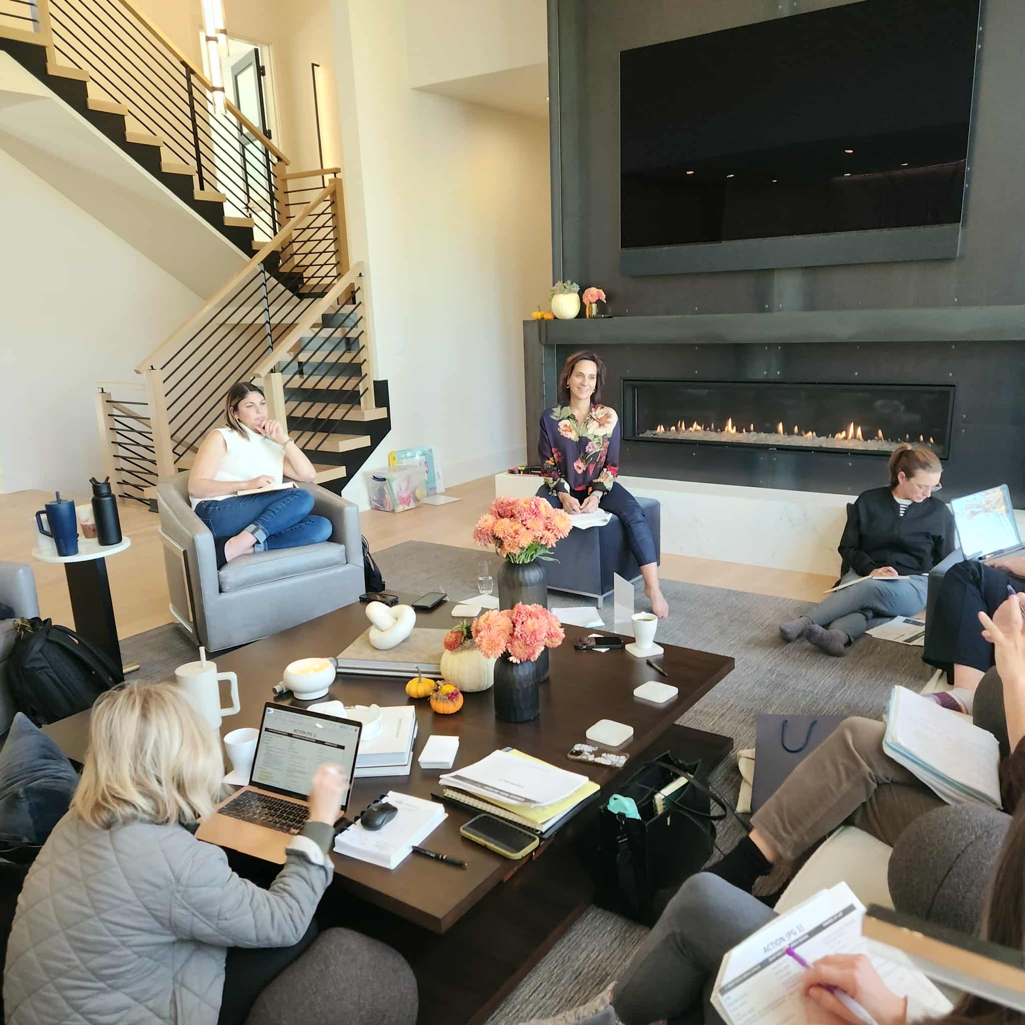 Members of She Thinks Big Collective seated around a table in a modern living room, some working on laptops and reading papers, with a woman sitting on the floor, a fireplace, and staircase in the background, decorated with vases and flowers.
