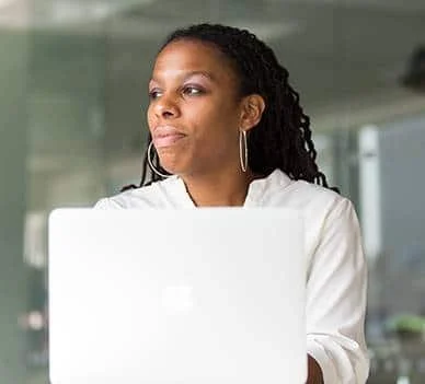 A woman with curly hair wearing hoop earrings and a white top, sitting at a desk with a laptop.