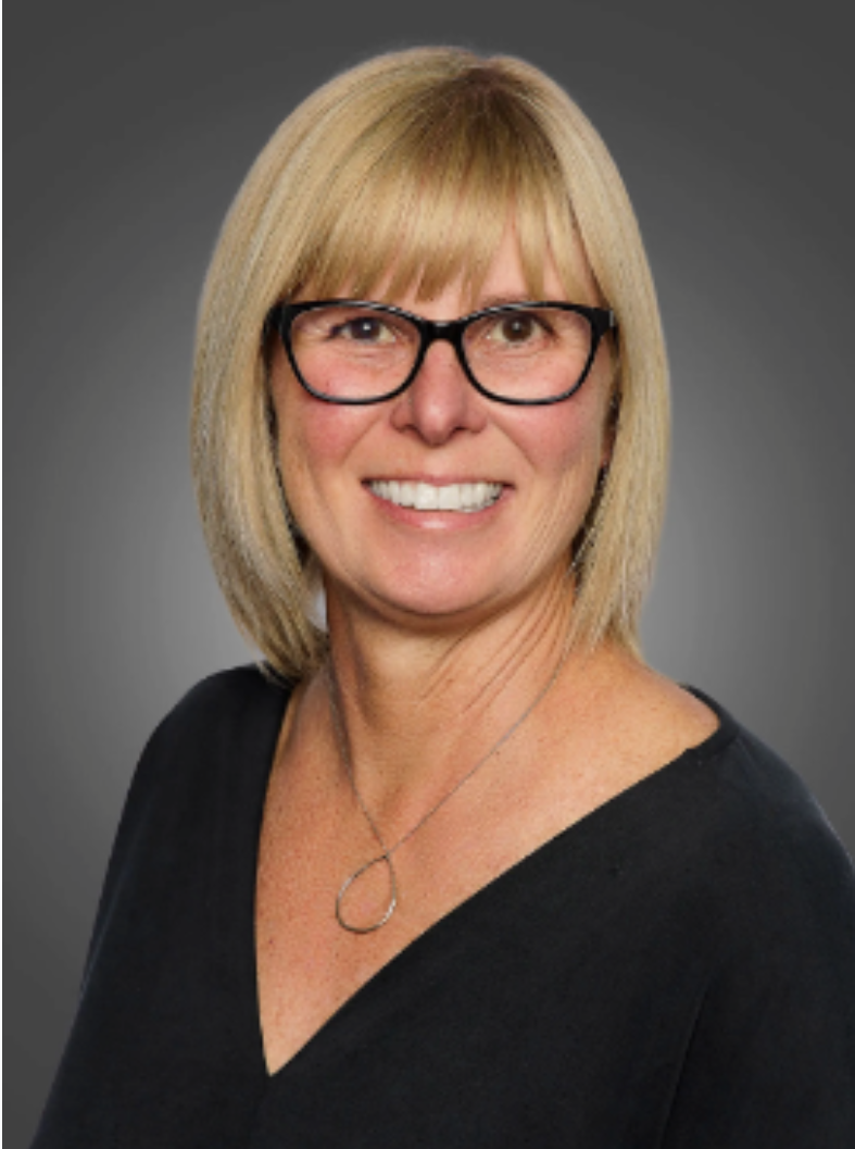 A middle-aged woman with blonde hair and glasses, wearing a black top and a silver necklace, smiling against a gray background.