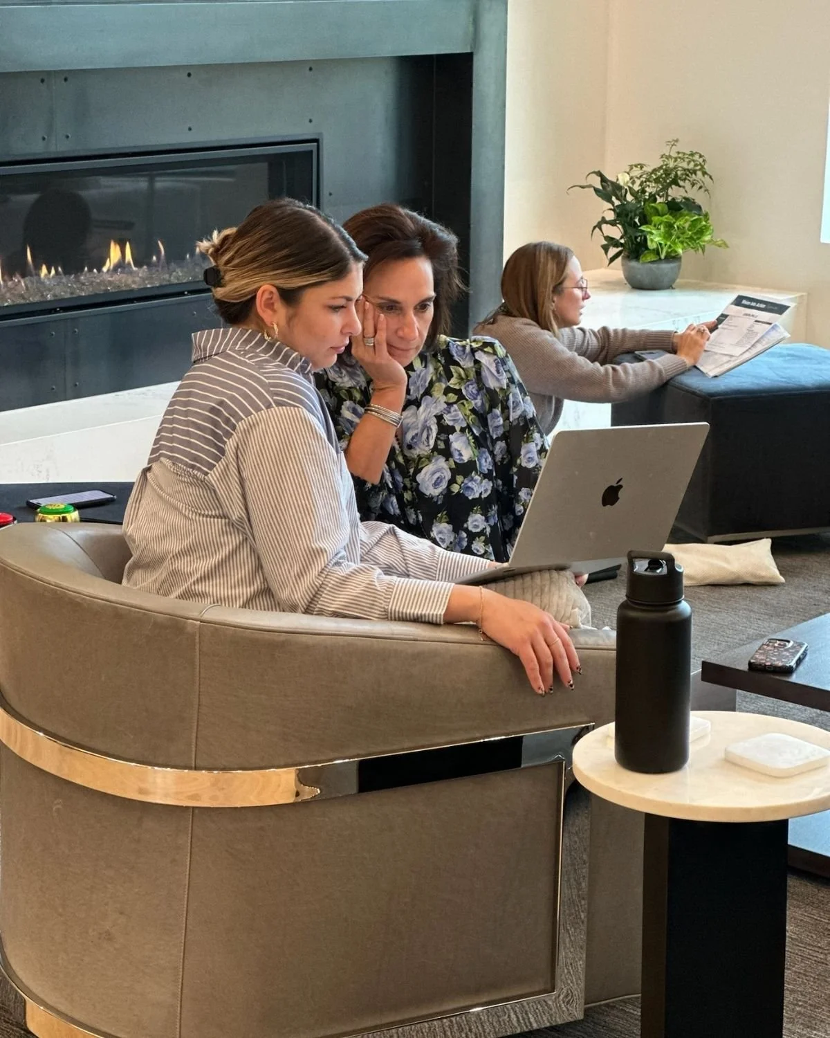Andrea Liebross coaching a client. Women sitting on a curved sofa, working on laptops, with a fireplace and potted plant in the background.