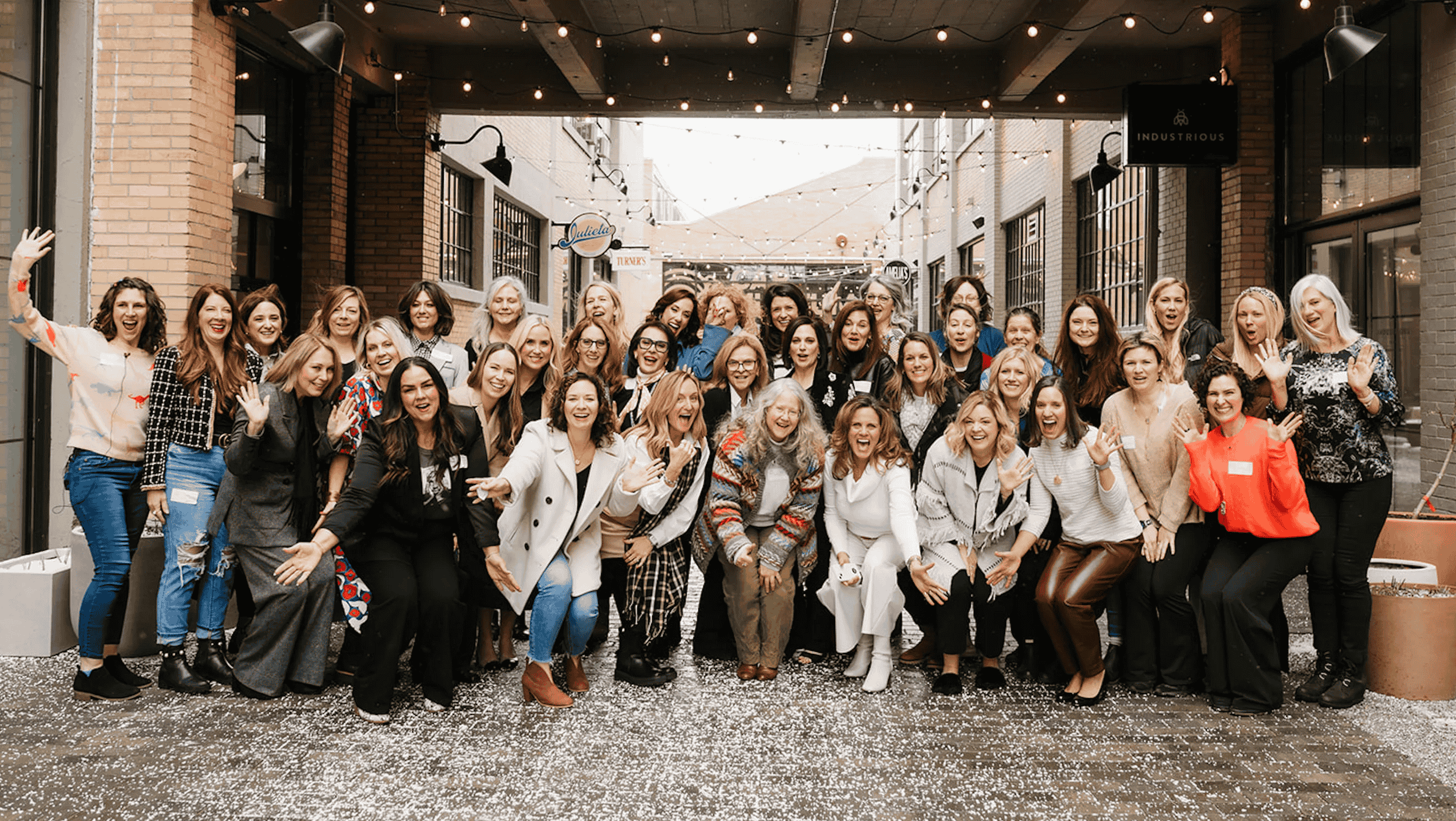 Collective members gathered for an in-person retreat. A large group of women smiling and waving at a gathering in an indoor-outdoor space decorated with string lights and confetti on the floor.