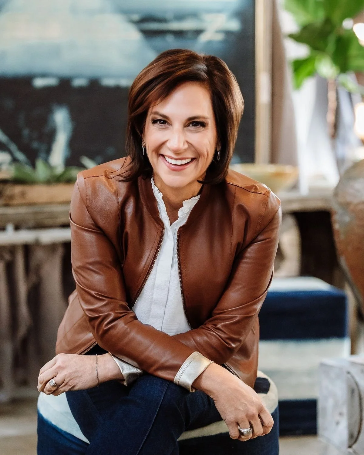 Andrea Liebross, a woman with short brown hair, wearing a brown leather jacket over a white shirt, smiling in a cozy indoor space with plants and decor in the background.