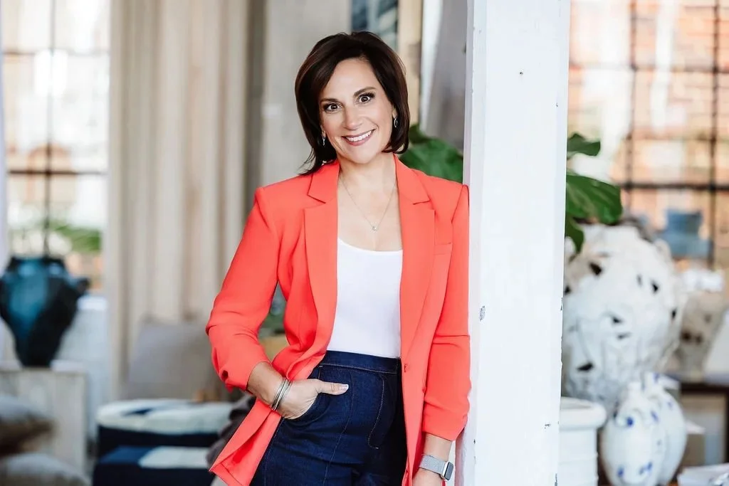 A woman with dark brown hair wearing a coral blazer and white top standing in a stylish indoor space with plants and decorative items.