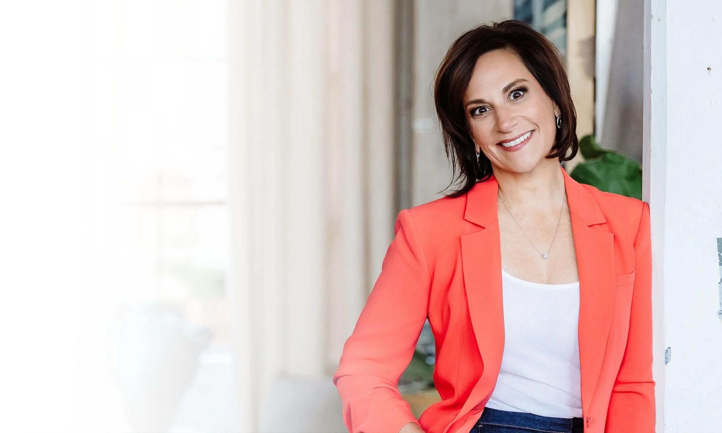 Andrea Liebross, a woman with short brown hair wearing a coral blazer and white top is smiling and leaning against a wall in an indoor setting.