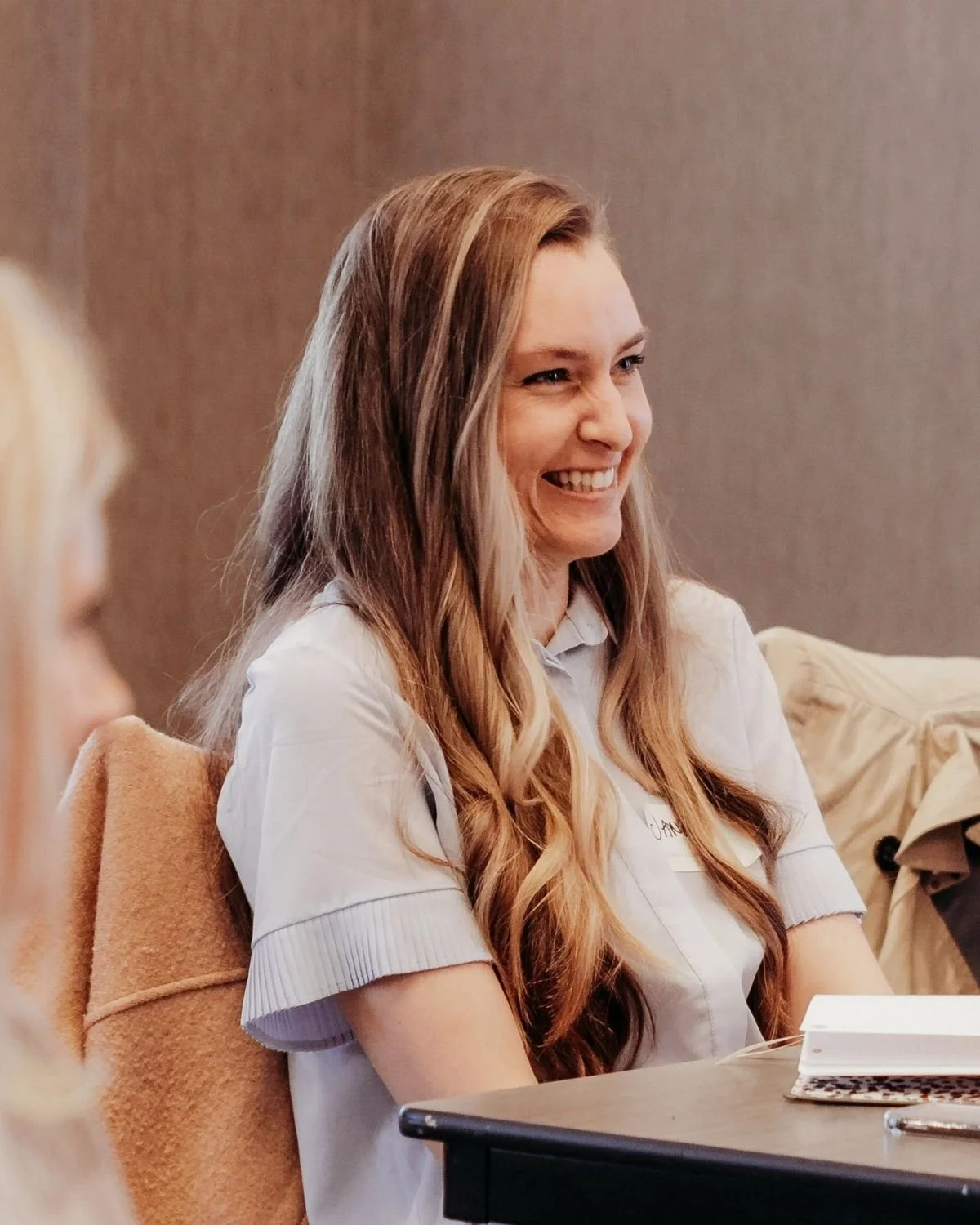 Young woman with long, wavy reddish-blonde hair smiling while sitting at a table during a meeting or discussion.