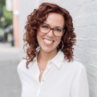 A woman with curly red hair and glasses smiling, wearing a white shirt, outdoors in a bright setting.