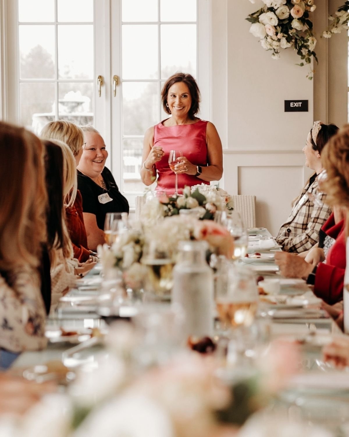 Andrea Liebross welcomes her guests to a She Thinks Big Collective dinner. She's standing, dressedin a red sleeveless dress giving a toast at a formal gathering, surrounded by seated guests, in a bright room with large windows and floral decorations.