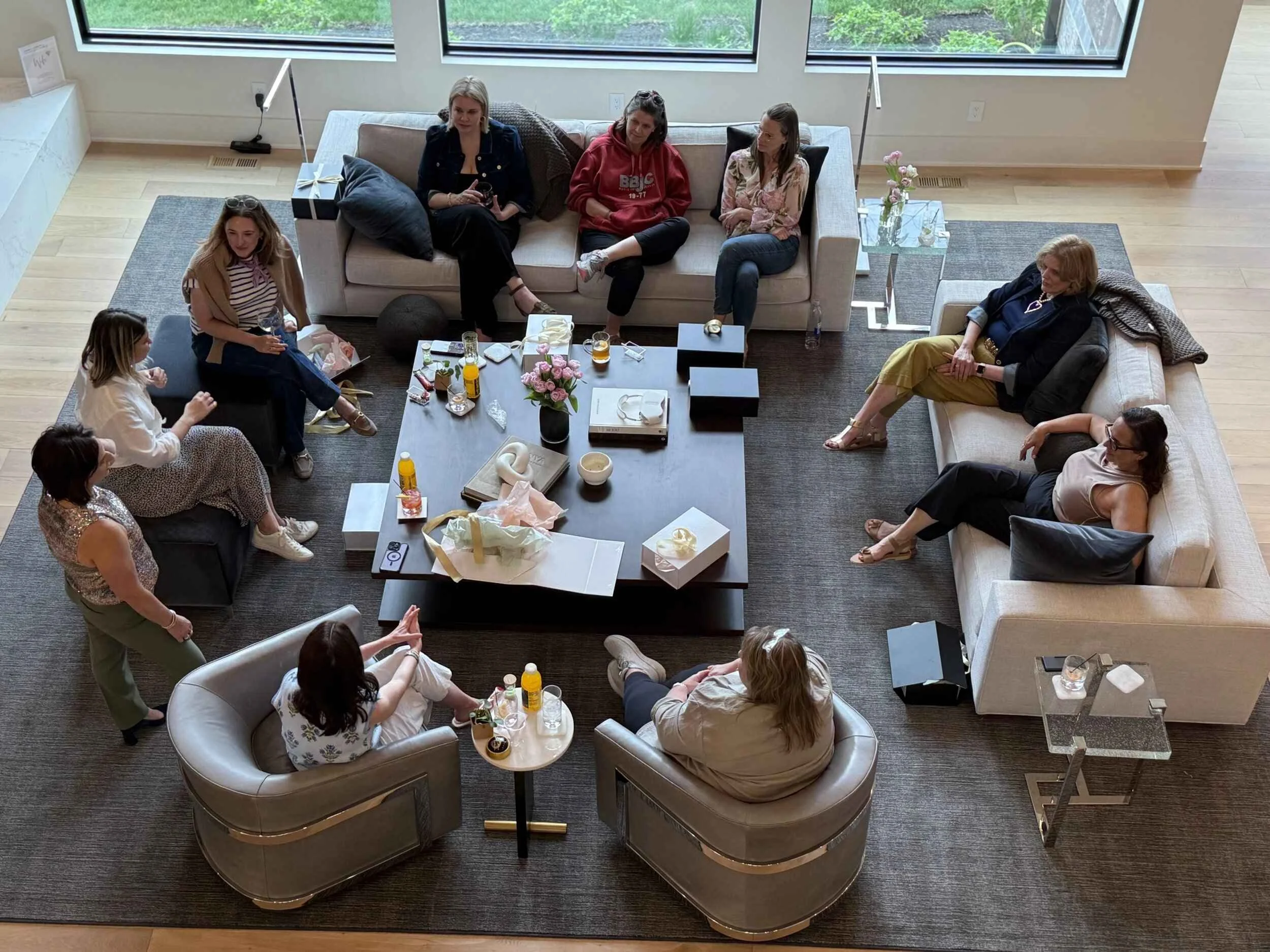 Women leaders in conversation during a Collective gathering. A group of women sitting and standing in a modern living room with large windows, couches, and a coffee table with flowers, drinks, and snacks.
