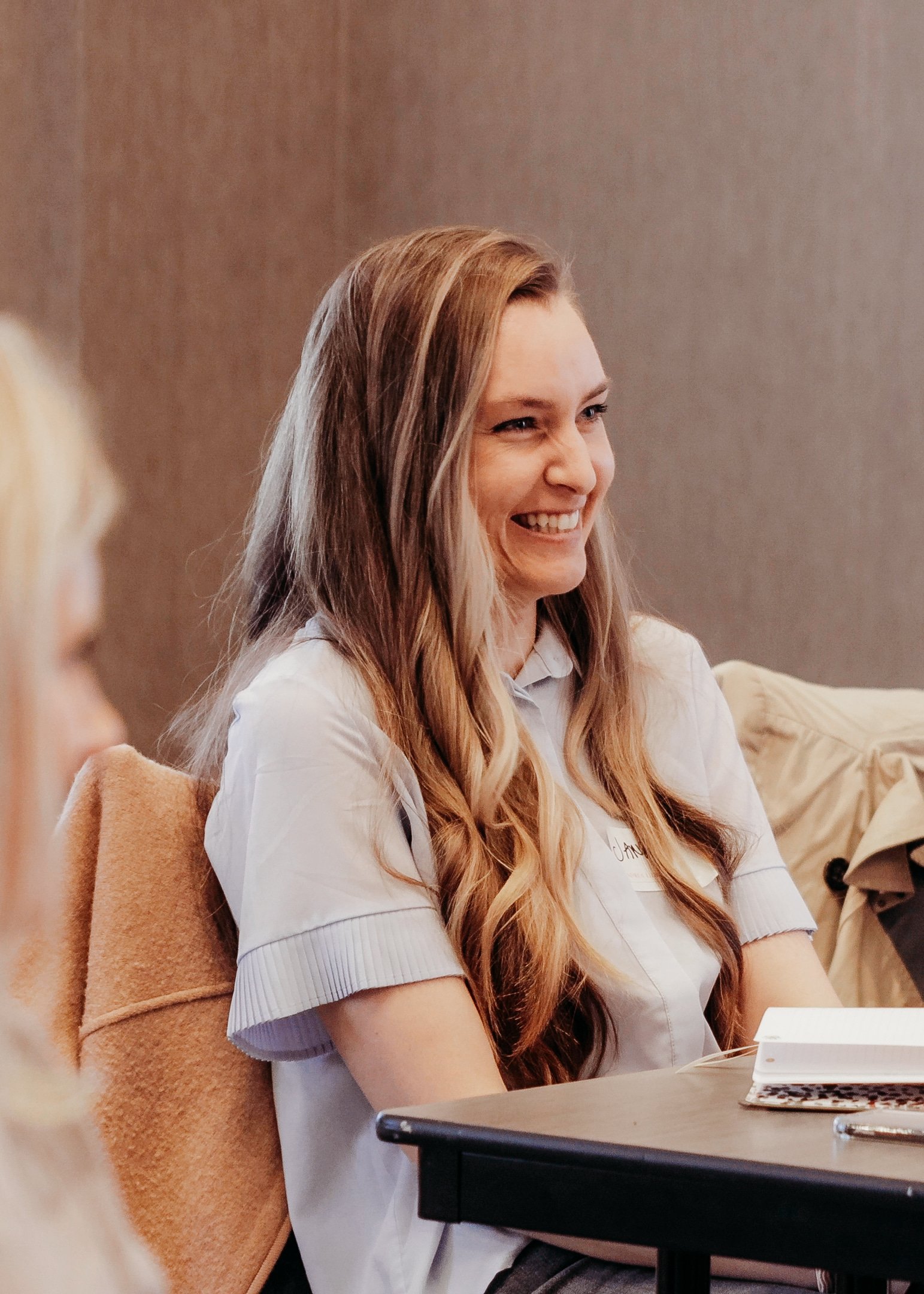 Young woman with long, wavy reddish-blonde hair smiling while sitting at a table during a meeting or discussion.