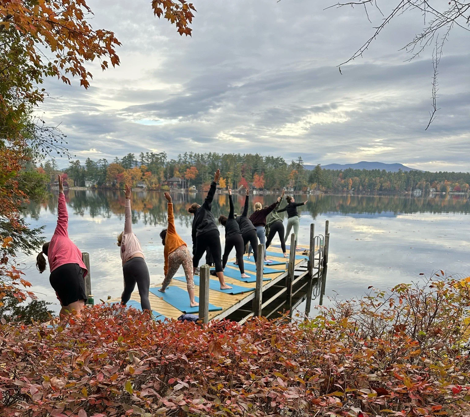 Women practicing yoga on a lakeside dock surrounded by autumn trees during a past She Thinks Big retreat.