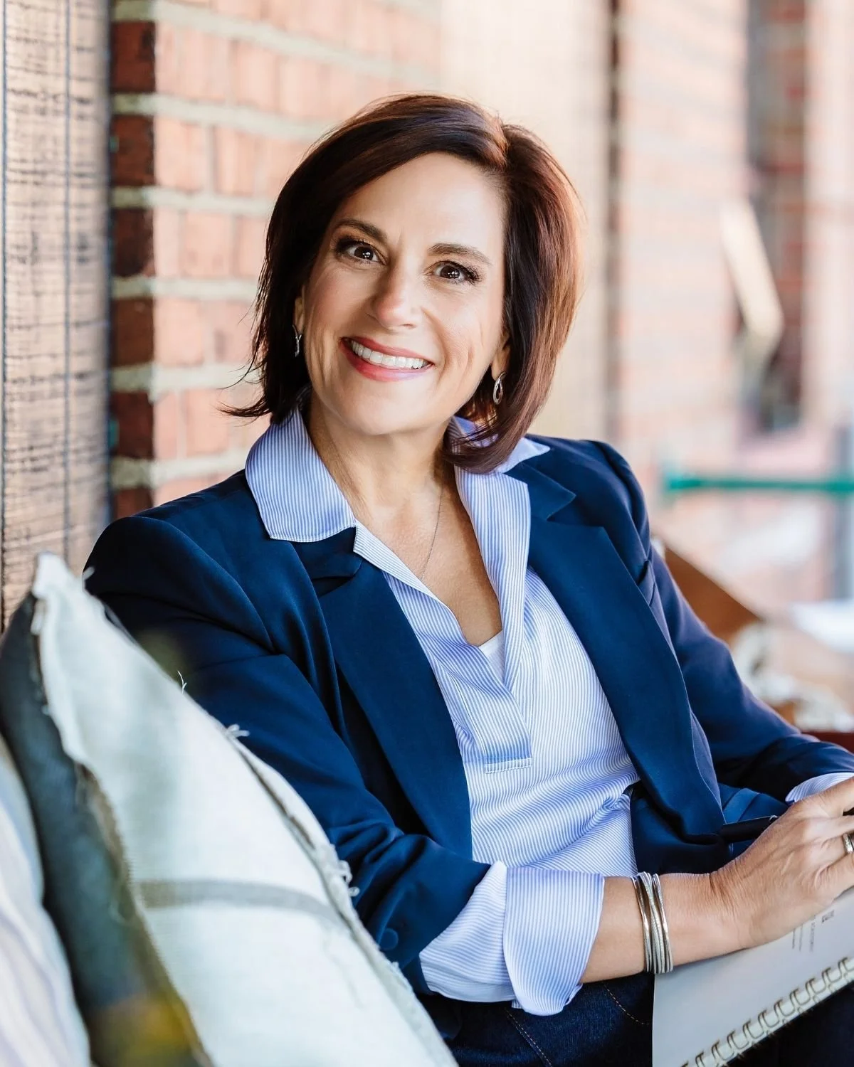 Andrea Liebross, a female with shoulder-length brown hair smiling, wearing a navy blazer and striped shirt, sitting outdoors against a brick wall.