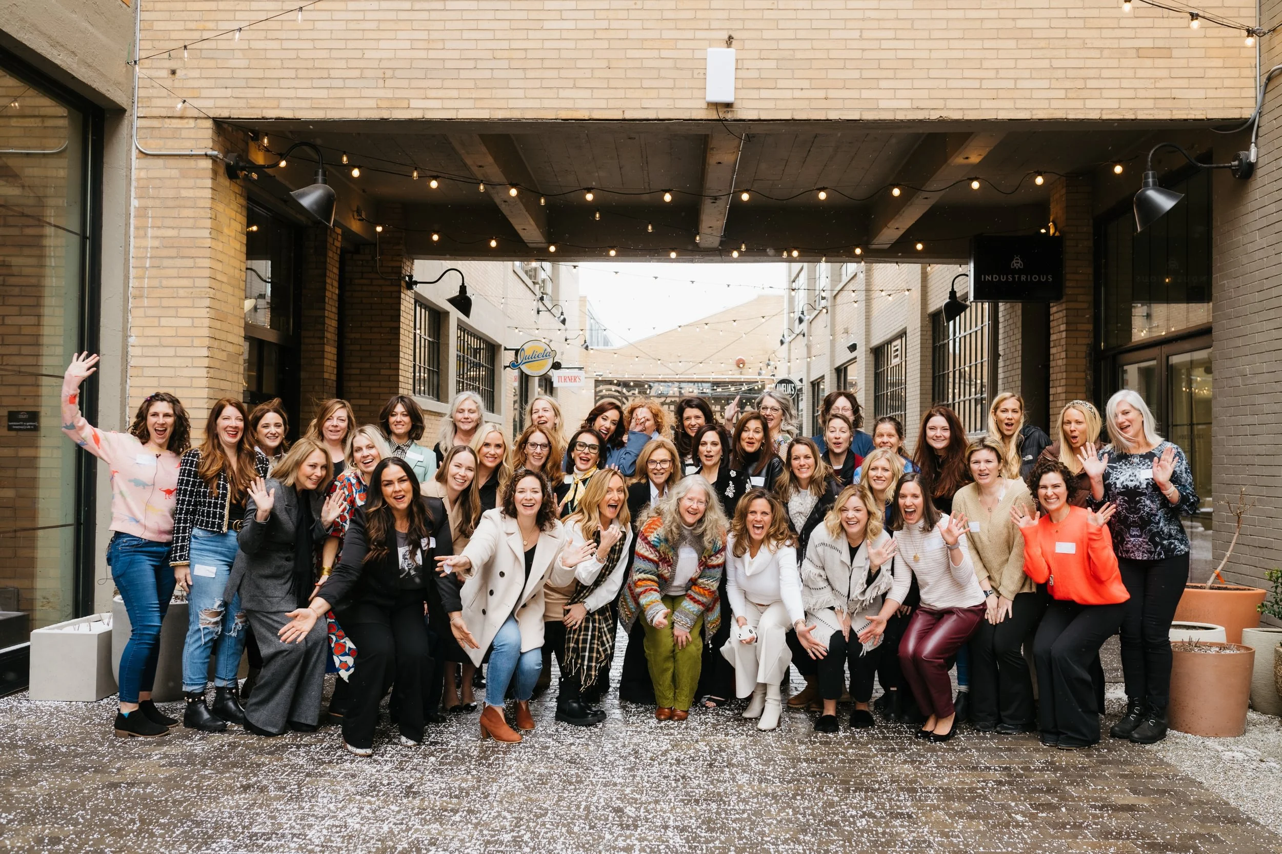 Group of women celebrating and smiling in an outdoor urban space with string lights overhead and brick walls.