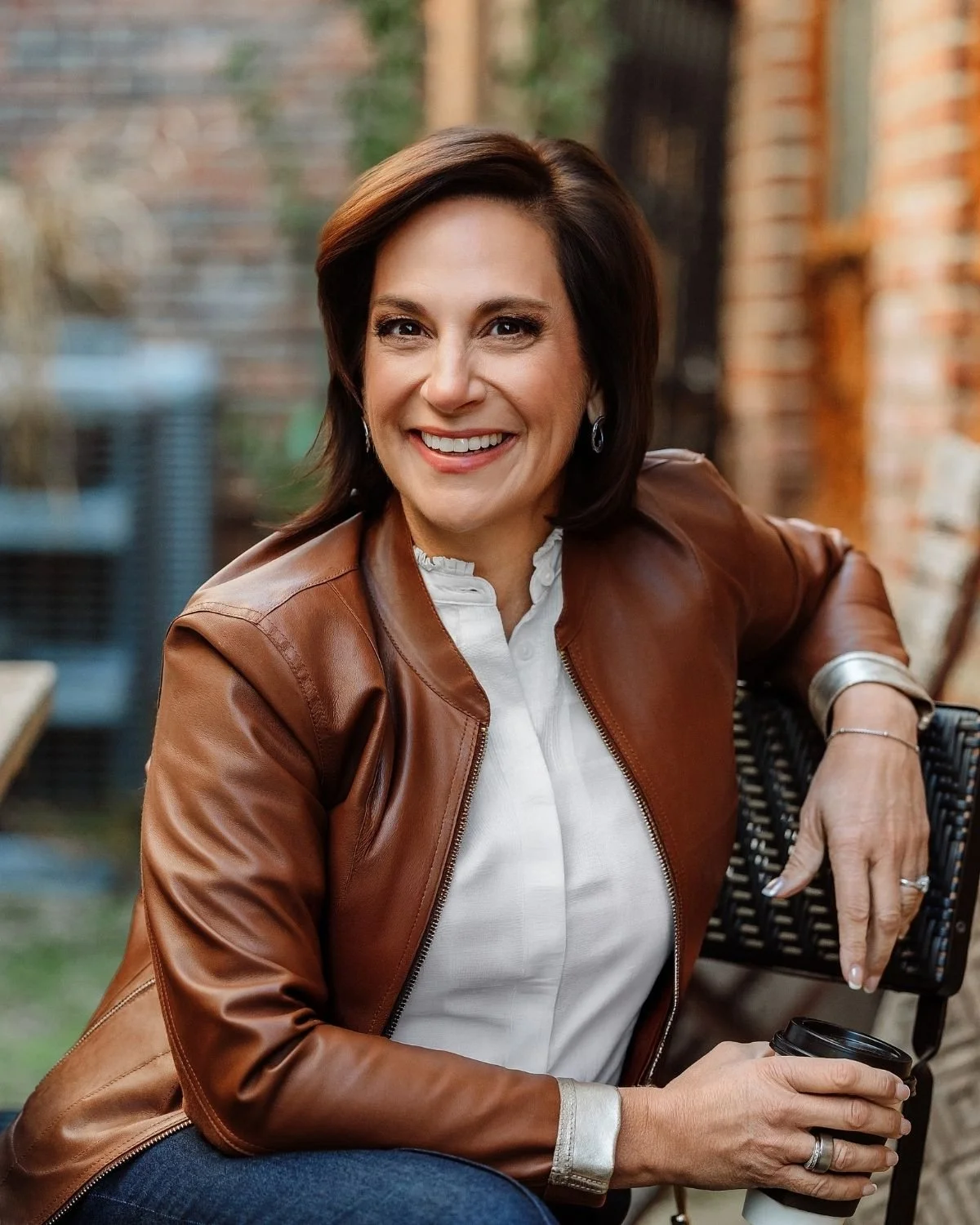 A woman, Andrea Liebross with shoulder-length dark brown hair smiling, wearing a brown leather jacket over a white blouse, sitting outdoors with a brick wall in the background, holding a black coffee cup.