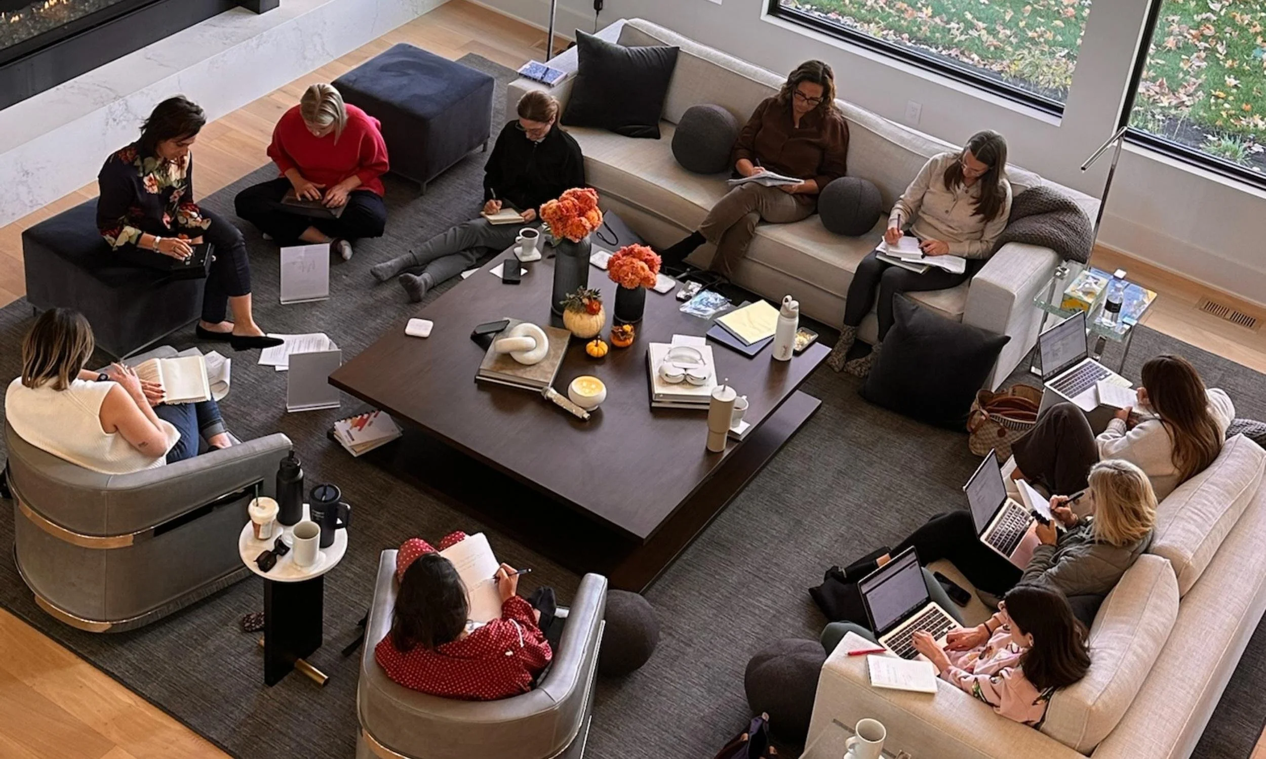 A group of nine business women, gathered for a She Thinks Big Collective event, sitting in a living room with large windows, working on laptops, notebooks, and tablets, surrounded by coffee cups, flowers, and various personal items.