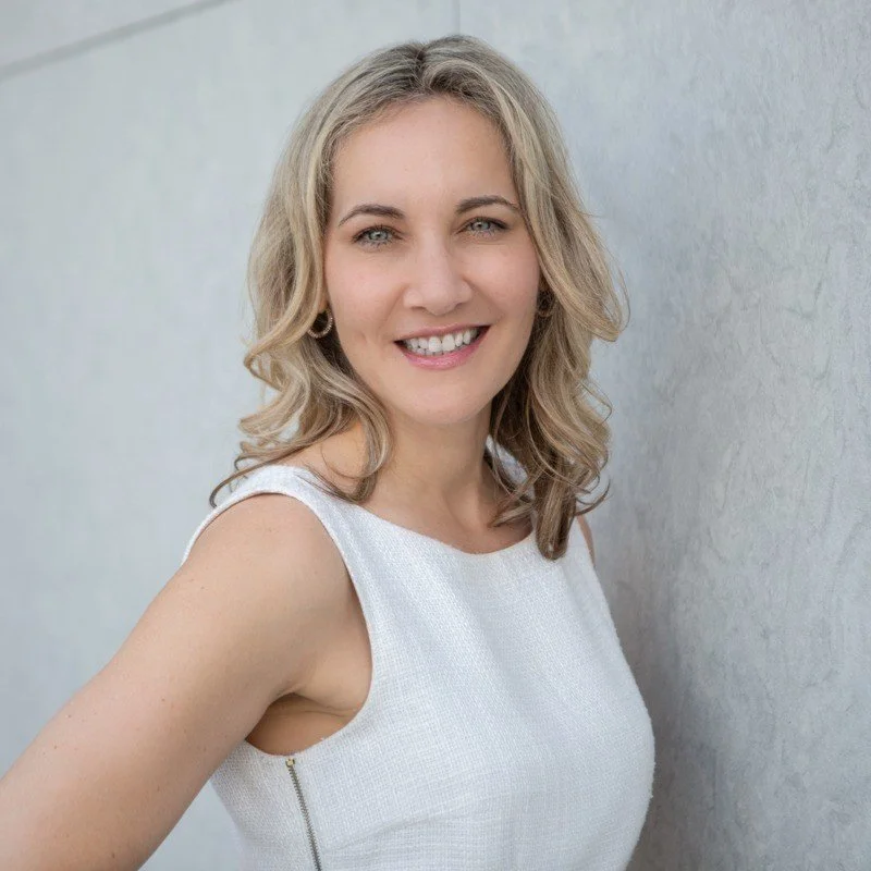 A woman with blonde, curly hair smiling and wearing a sleeveless white top, standing in front of a plain wall.