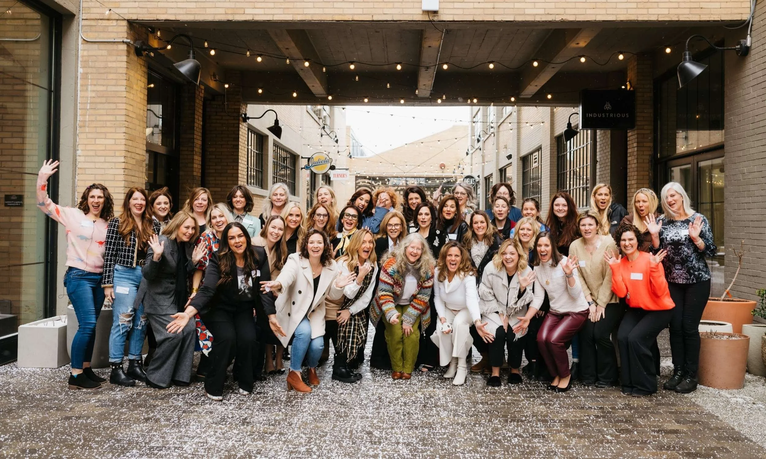 A large group of women gathered for a Shel Thinks Big event, outdoors beneath string lights, smiling and waving, in a brick alleyway with potted plants and a confetti-like ground cover.