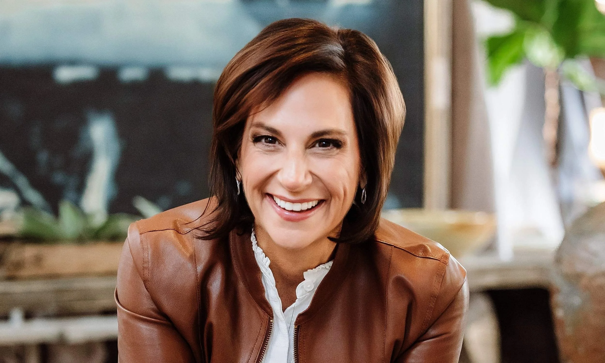 Andrea Liebross, a woman with short brown hair, wearing a brown leather jacket over a white shirt, smiling in a cozy indoor space with plants and decor in the background.