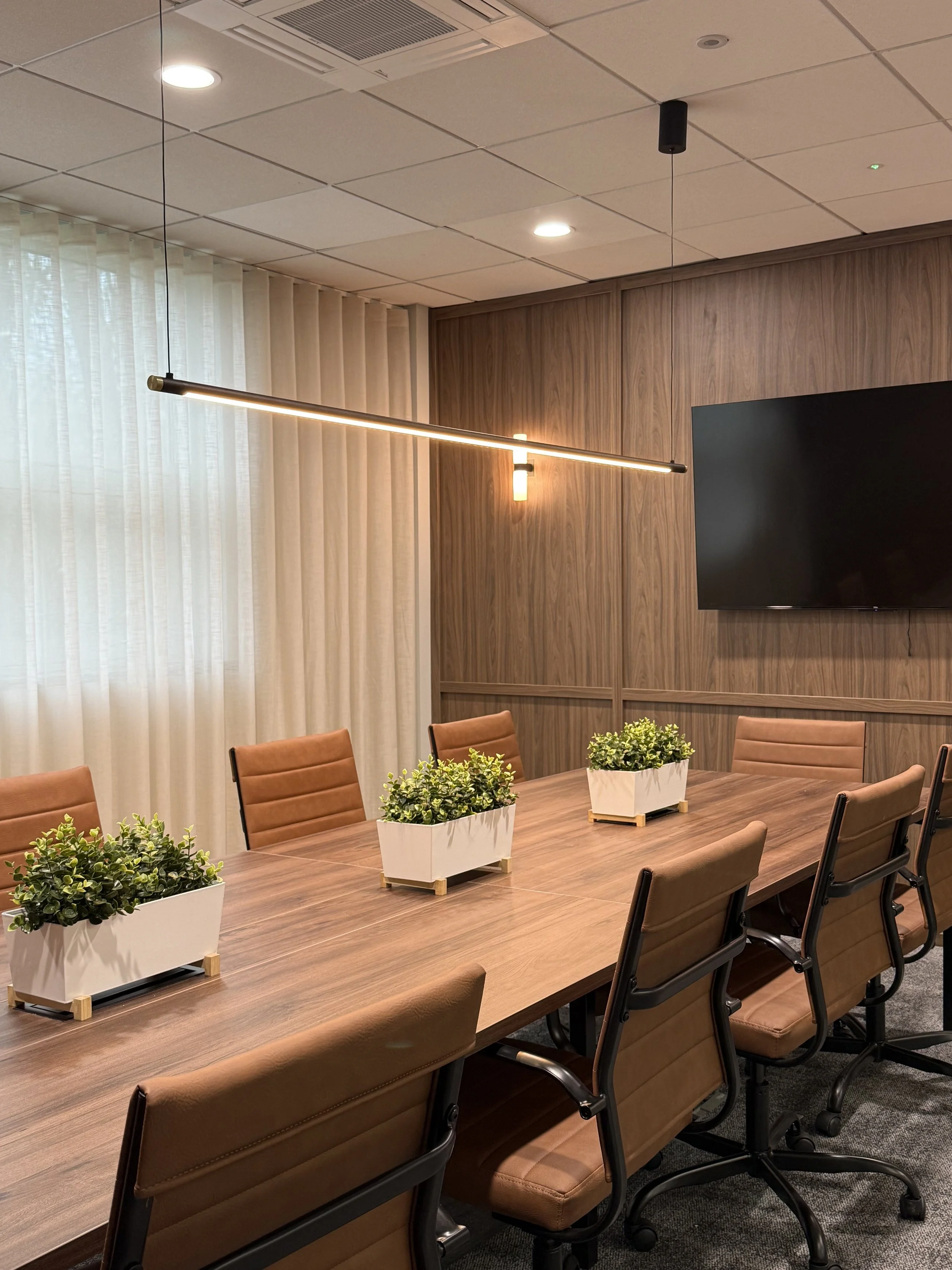 A modern conference room with a long wooden table, brown office chairs, potted plants, a large wall-mounted TV, curtains, and ceiling lighting.