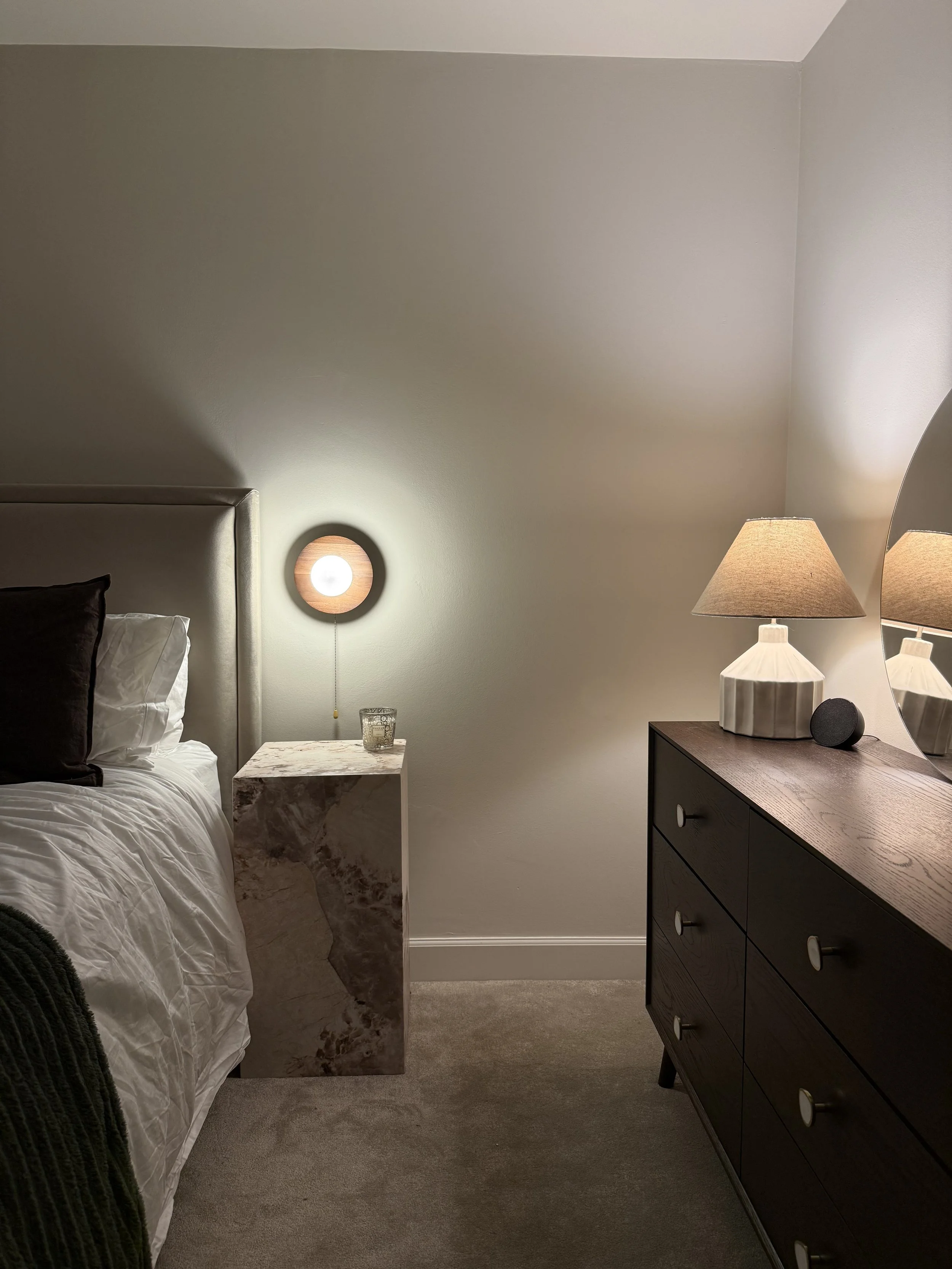 A bedroom corner with a bed, a marble nightstand, a wall-mounted light, and a dark wooden dresser with table lamps and a mirror.