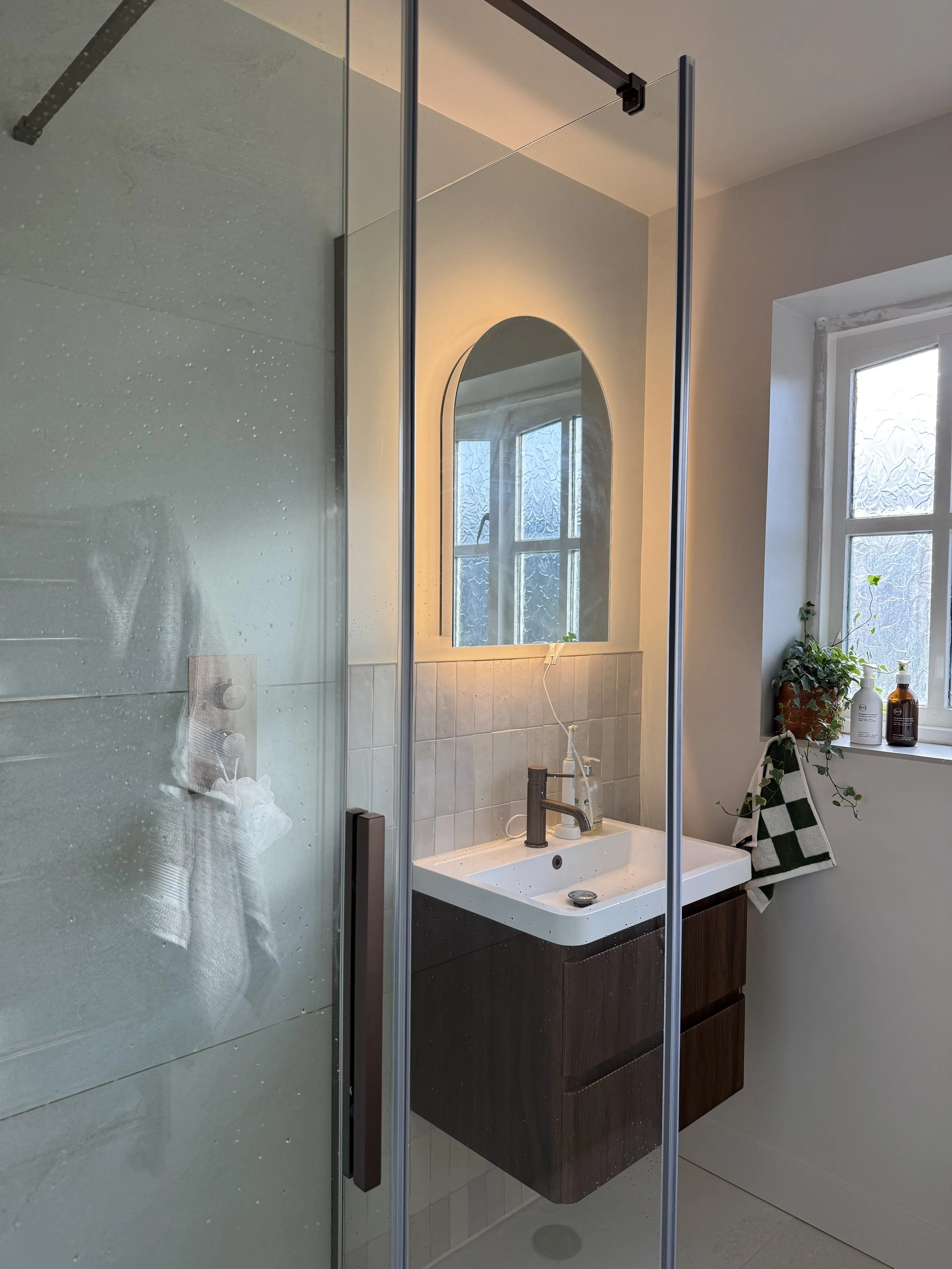 A modern bathroom with a shower enclosure, a white ceramic sink with a dark wooden cabinet, a round mirror with backlighting, a window with frosted glass, and a potted plant on the windowsill.