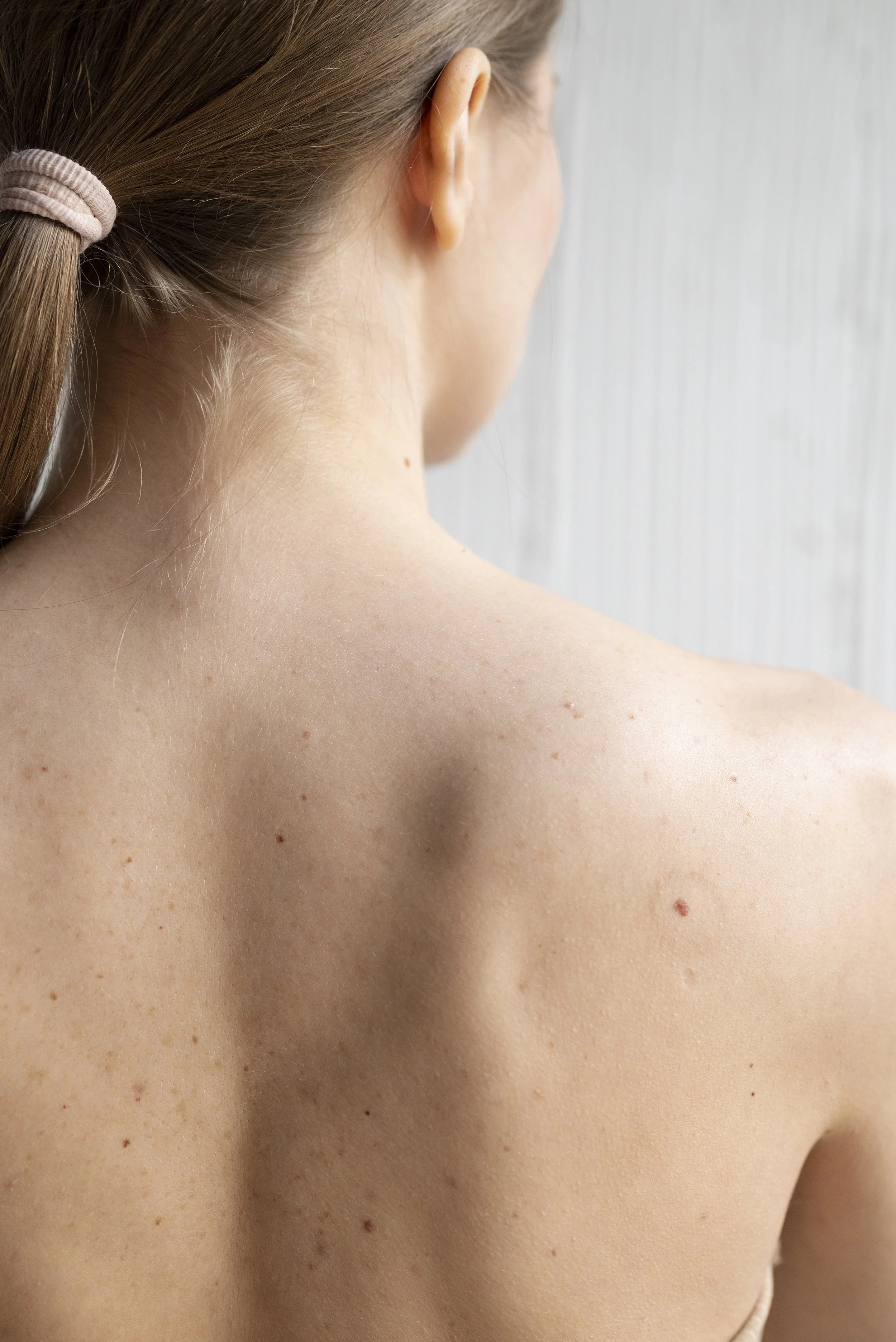 Close-up of a woman's back and shoulder, showing her skin and hair tied with a pink scrunchie, against a light-colored wall.