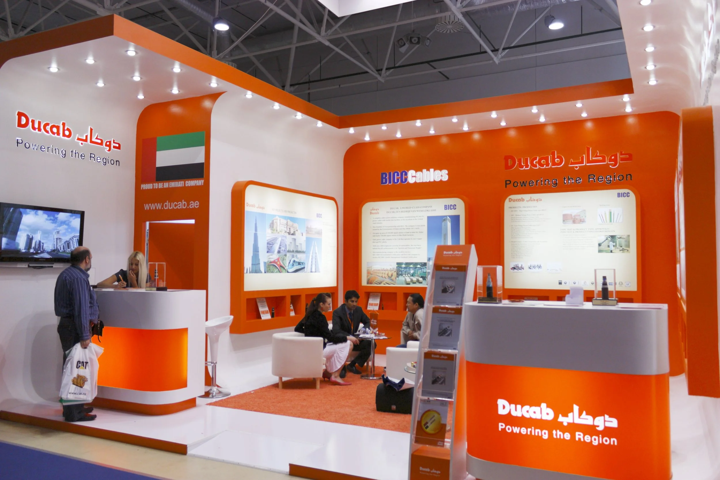 An exhibition booth for Ducab with orange and white color scheme, featuring informational displays, a reception desk, and a small seating area with four people engaged in conversation.